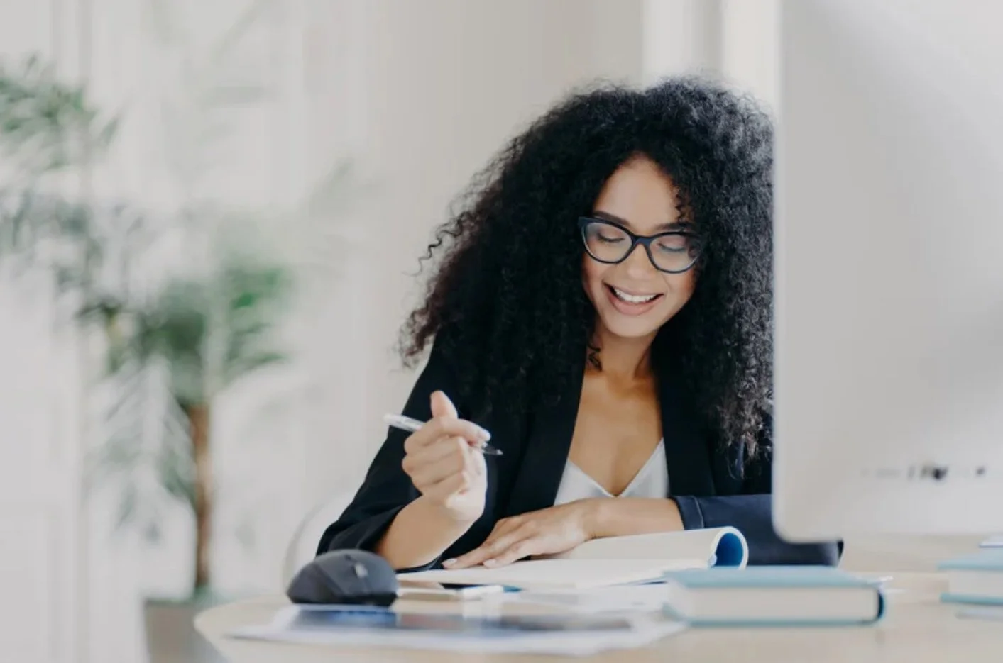 A woman with curly hair, glasses, and a black blazer smiling and writing at a desk with books and a computer mouse.