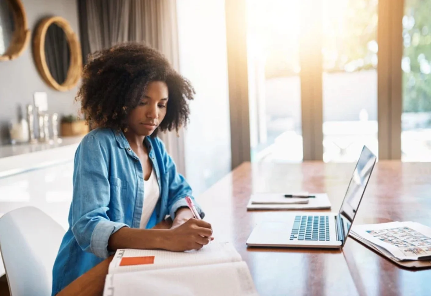A woman with curly hair writing in a notebook at a wooden table with an open laptop and papers, sunlight streaming through large windows in the background.
