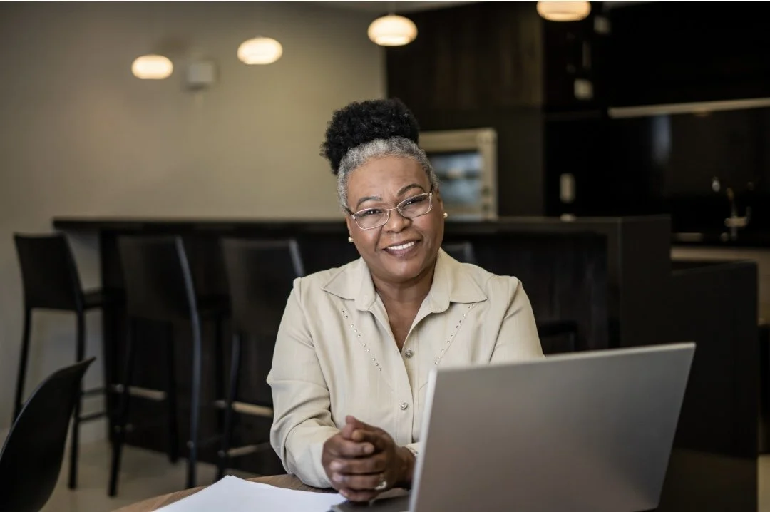 An older woman with glasses and gray hair styled in a bun, sitting at a table with a laptop in a modern kitchen or office space.