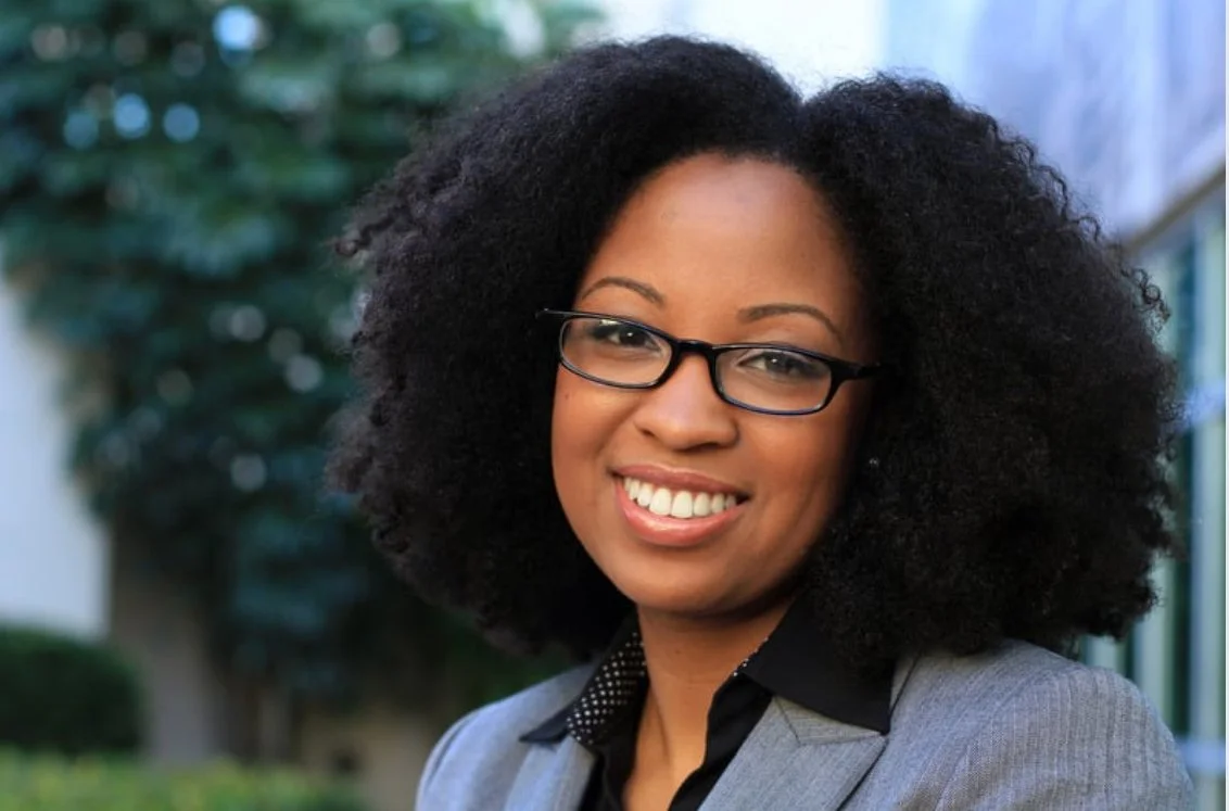 A smiling woman with black glasses and curly black hair, wearing a gray blazer and black shirt, outdoors with greenery in the background.