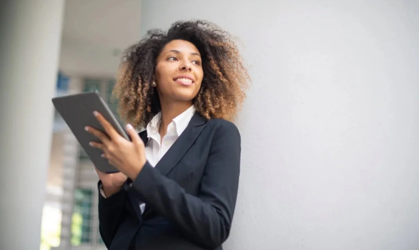 A young woman with curly hair, wearing a black blazer and white shirt, holding a tablet, standing outdoors near a white wall, looking off to the side with a slight smile.