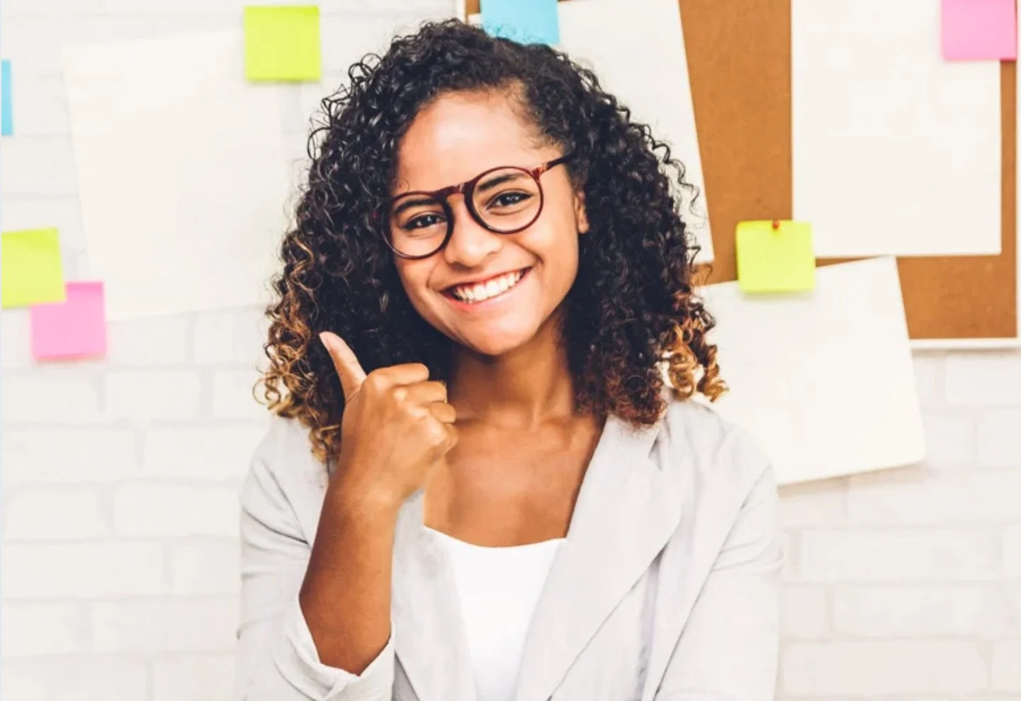 Smiling woman with curly hair and glasses pointing to her head in an office with colorful sticky notes on the wall.
