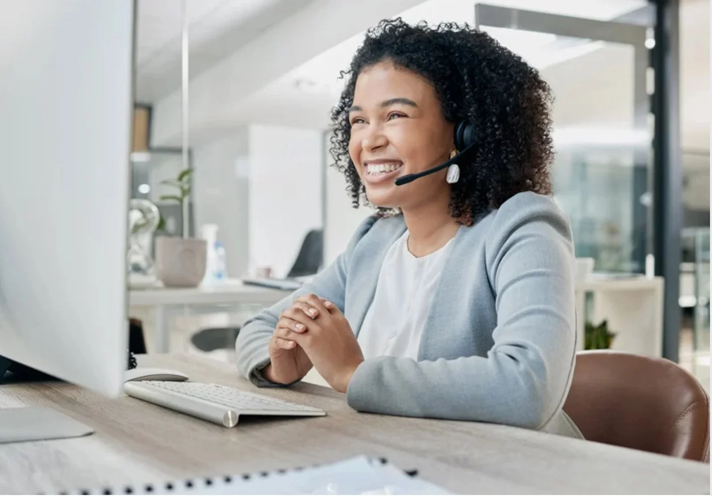 A woman with curly hair smiling while wearing a headset at her desk in an office setting, with a computer, keyboard, and office decor in the background.
