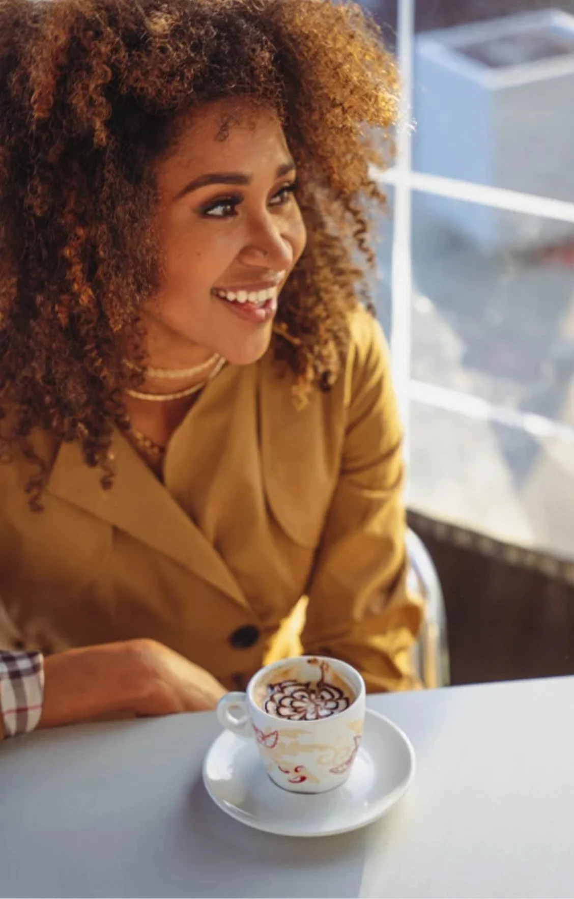 A woman with curly hair smiling in a café, sitting at a table with a cup of coffee in front of her.