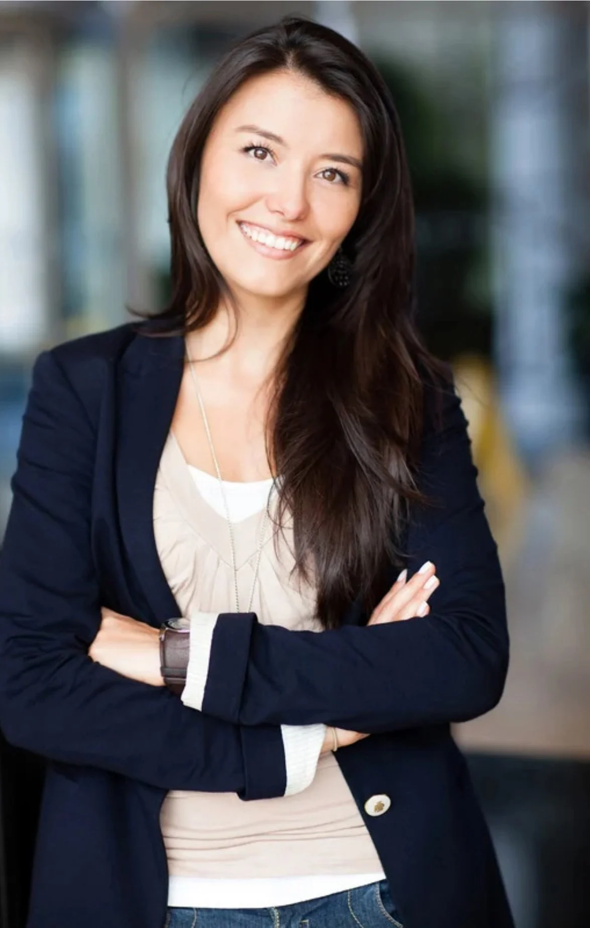 A smiling woman with long dark hair, wearing a navy blazer, white shirt, and jeans, standing outdoors with arms crossed.