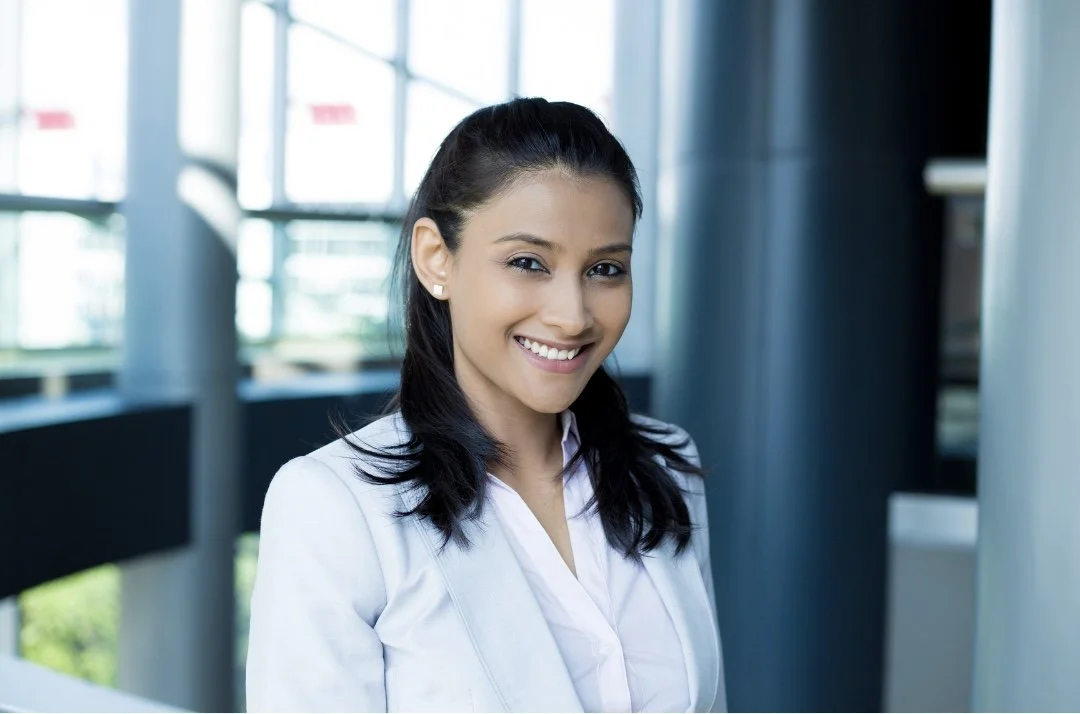 A young woman with dark hair, wearing a white blazer, smiling, standing in a modern office environment with large windows.