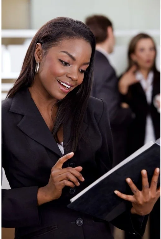 A smiling woman in professional attire looking at a notebook in an office setting with two blurred colleagues in the background.