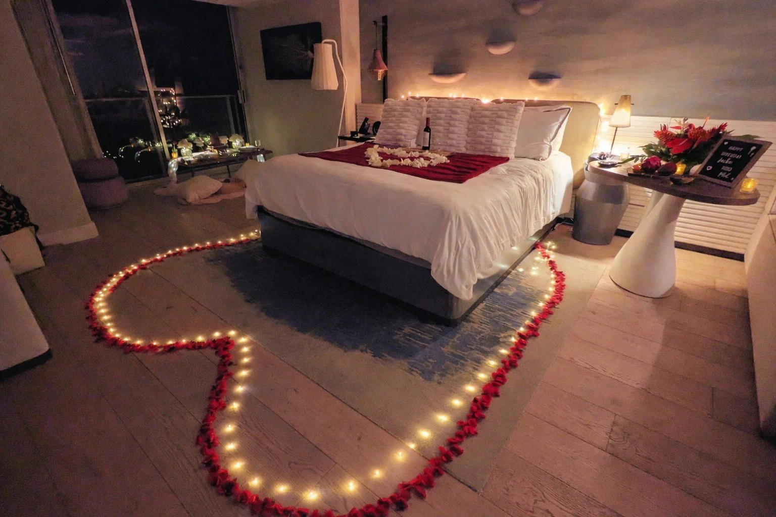 A bedroom decorated for a romantic occasion with a heart-shaped outline made of rose petals and string lights on the floor around the bed. The bed has pillows and a drink on it. There is a table with flowers, candles, and a sign that says "Happy Vale