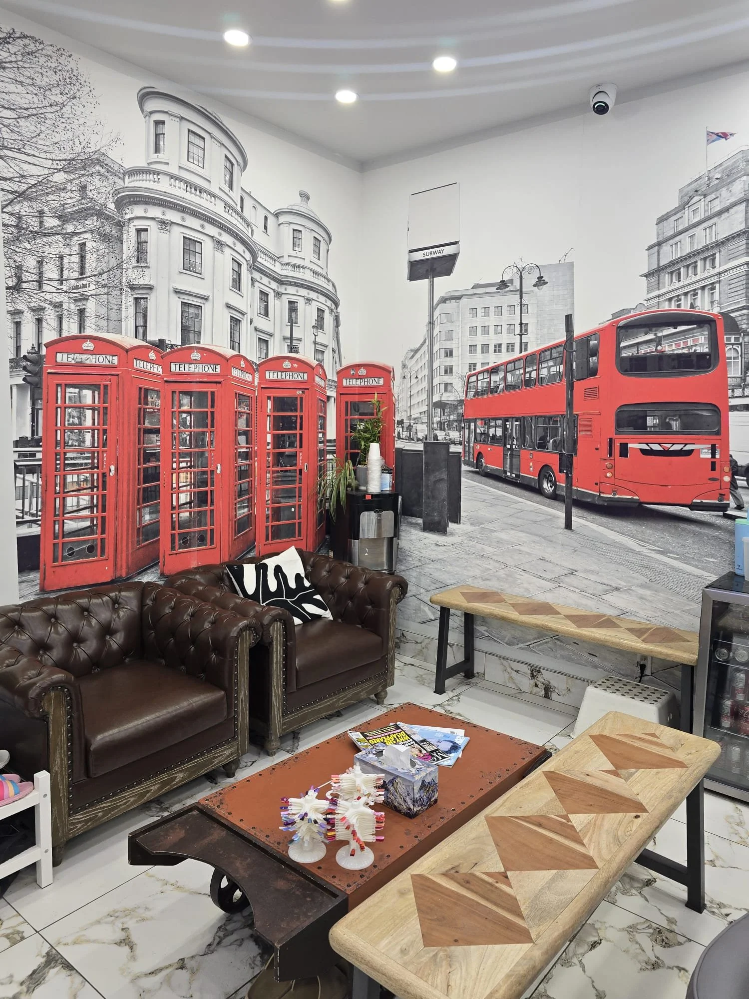 Indoor scene featuring a wall mural of London cityscape with red telephone booths and a double-decker bus. The room is furnished with a brown leather couch, wooden tables, and miscellaneous decor items, with a marble floor.