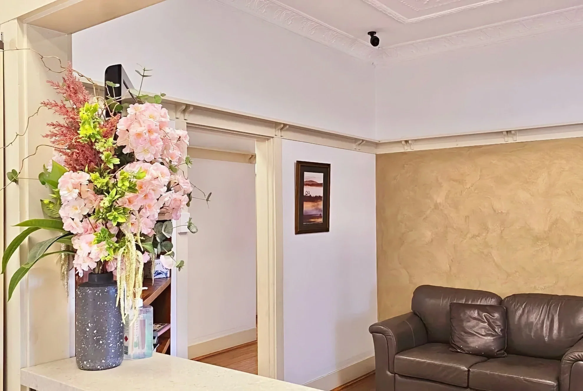 Living room corner with a leather sofa, a framed landscape picture on the wall, and a tall floral arrangement with pink flowers and green leaves on a table near a doorway.