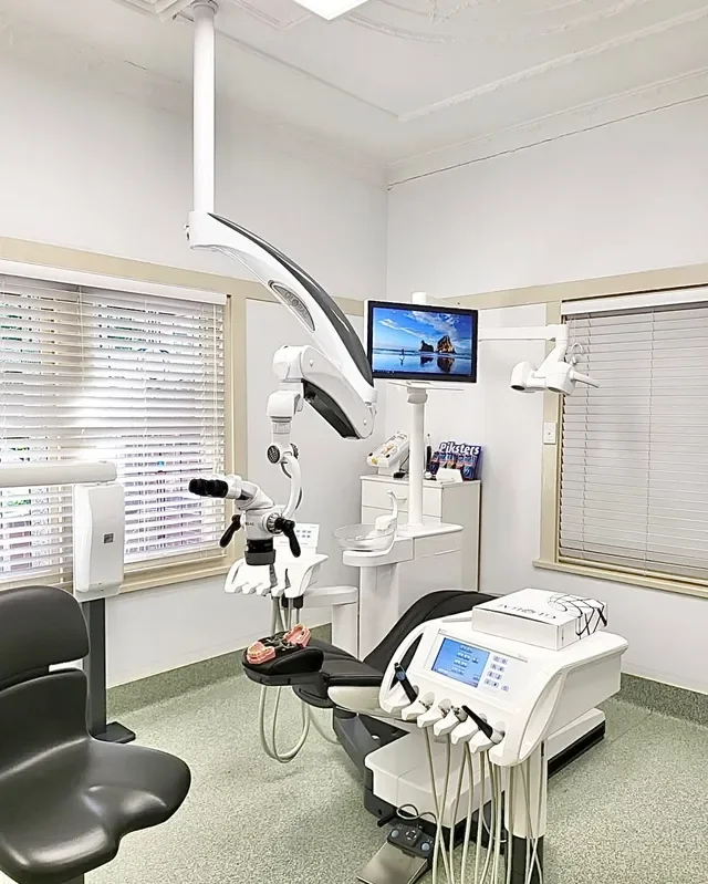 Dental examination room with dental chair, dental equipment, overhead lights, window blinds, and a monitor displaying a scenic beach view.