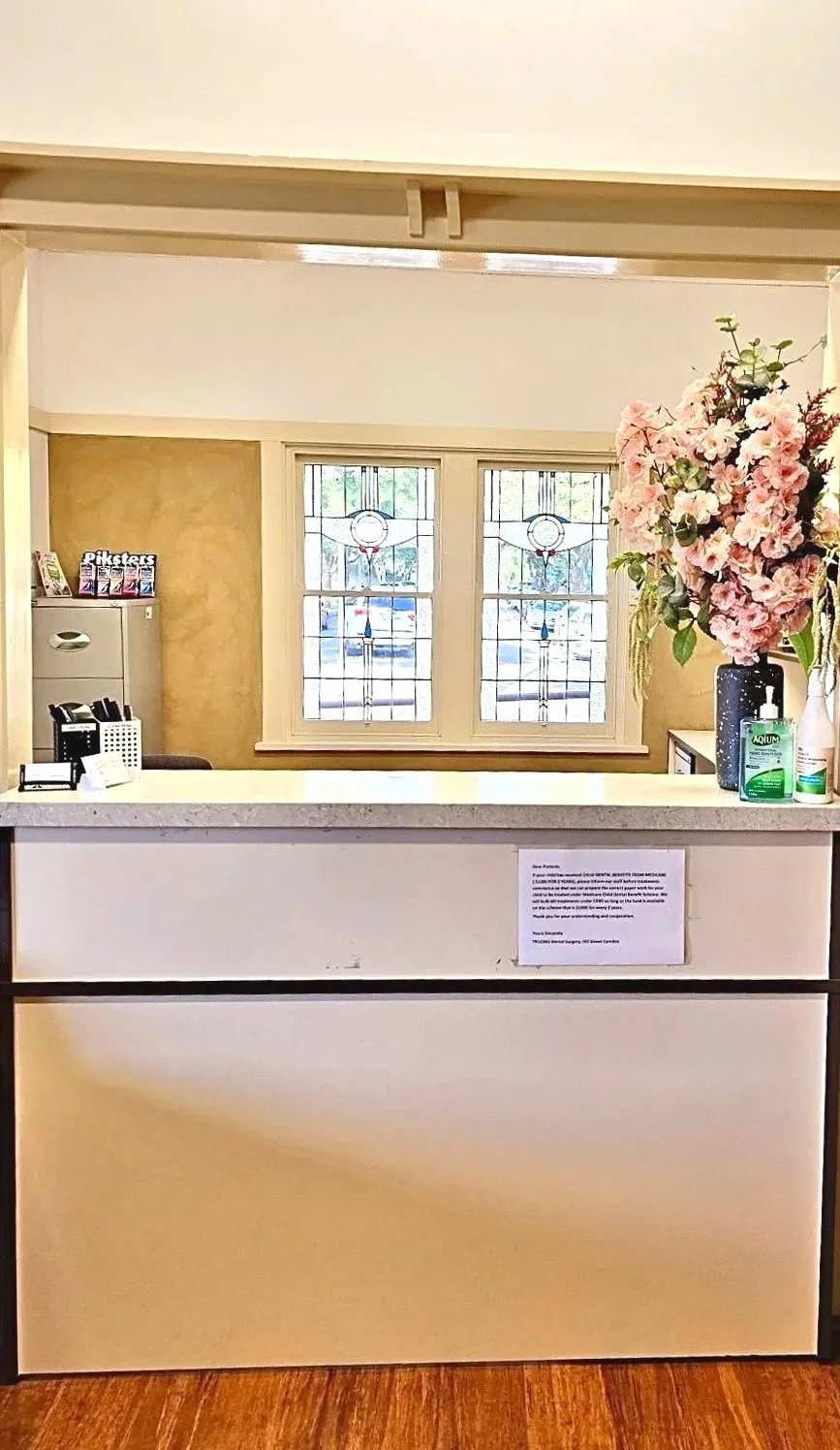 Reception desk with a sign, a vase of pink flowers, hand sanitizer, and a window with stained glass above.