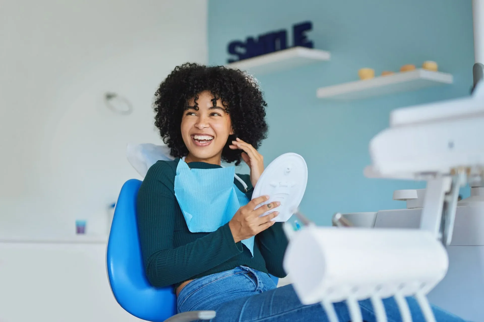 Young woman sitting in dental chair at dentist office, smiling and holding dental tray, wearing a blue bib and casual clothes.