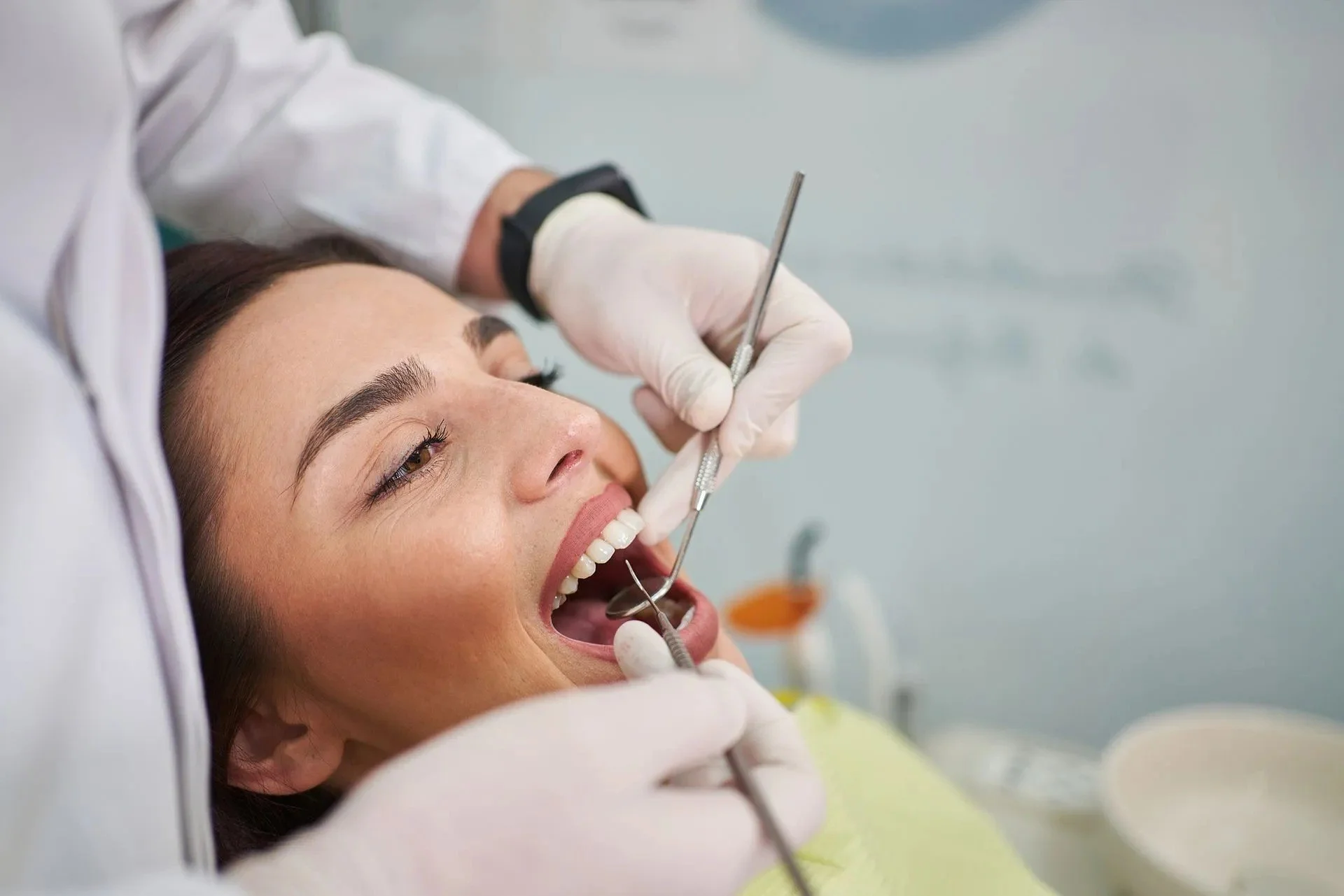 A woman lying back with mouth open during a dental visit, receiving dental care from a dental professional wearing gloves, holding dental tools.