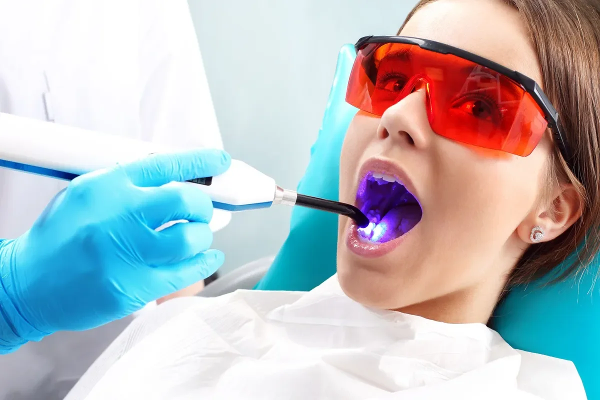 Dental patient wearing orange protective glasses undergoing a teeth whitening procedure with a dentist using a curing light. The patient has a white dental bib and is lying in a dental chair.