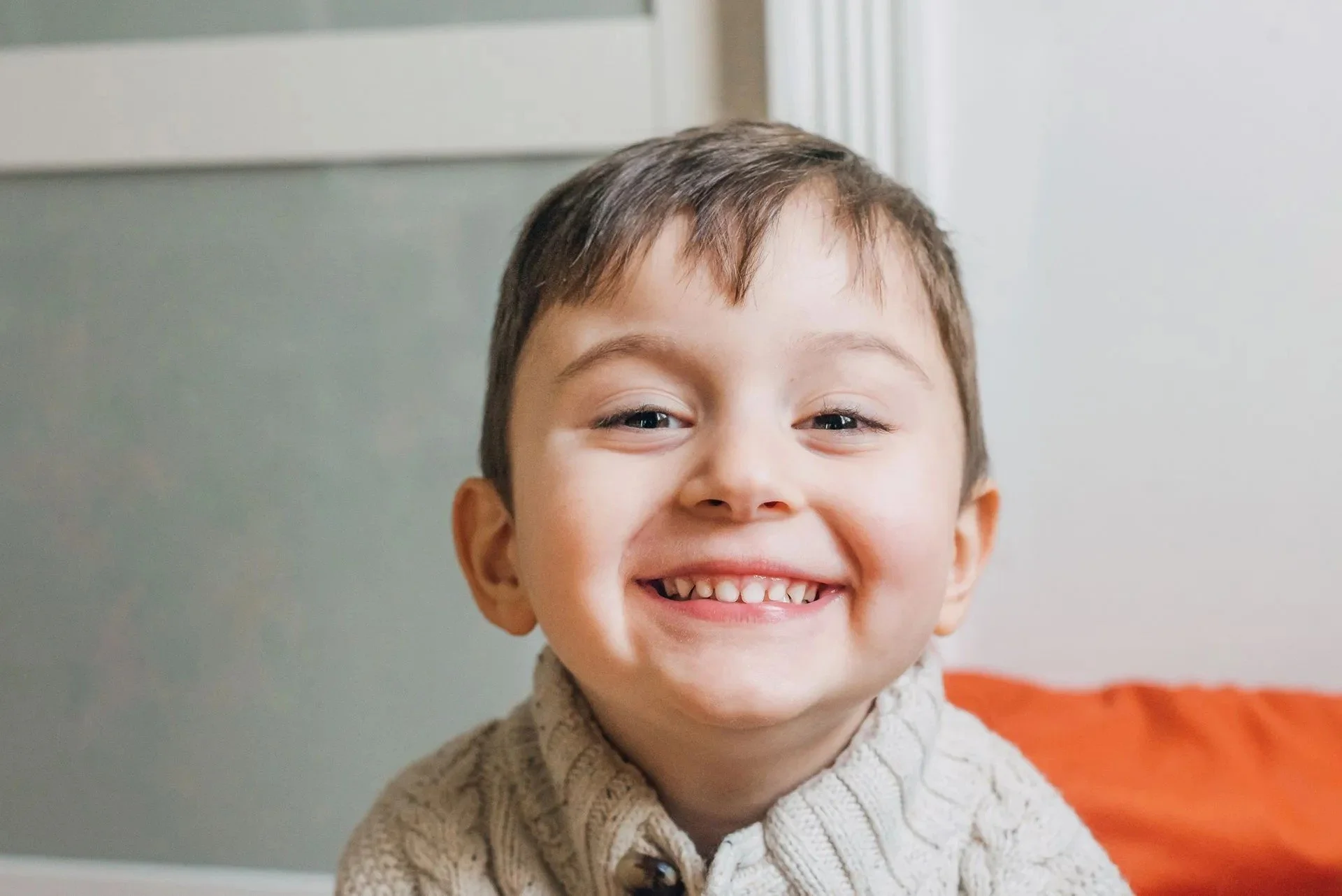 Close-up of a young boy with short brown hair and a big smile showing his teeth, wearing a beige knitted sweater, standing in front of a light-colored wall and window.