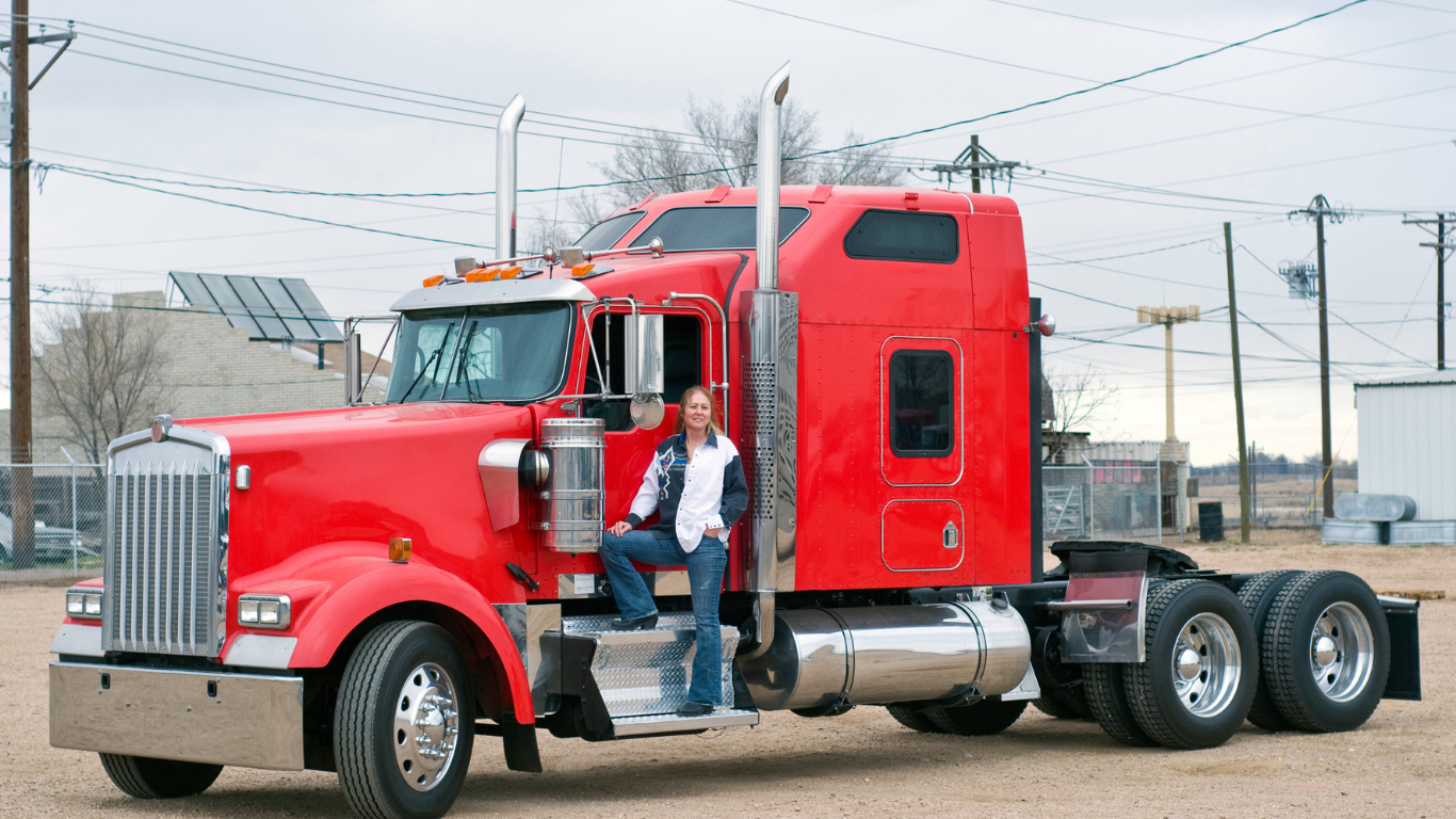 A woman sitting on the step of a large red semi-truck parked outdoors on a cloudy day.