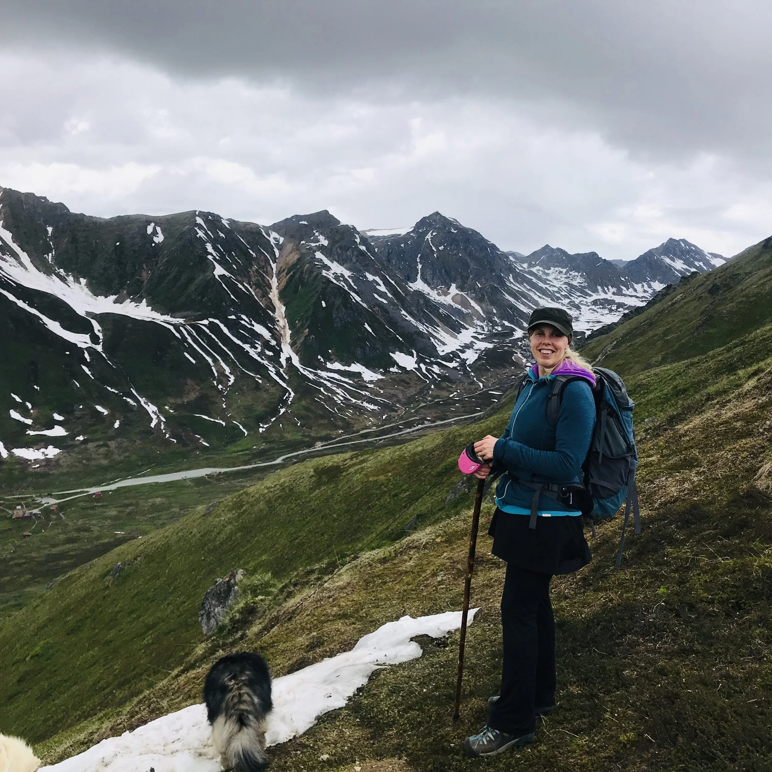A woman hiking in a mountainous landscape with snow patches, green hills, and cloudy sky, holding a hiking pole, wearing a backpack and outdoor clothing, with a dog nearby.