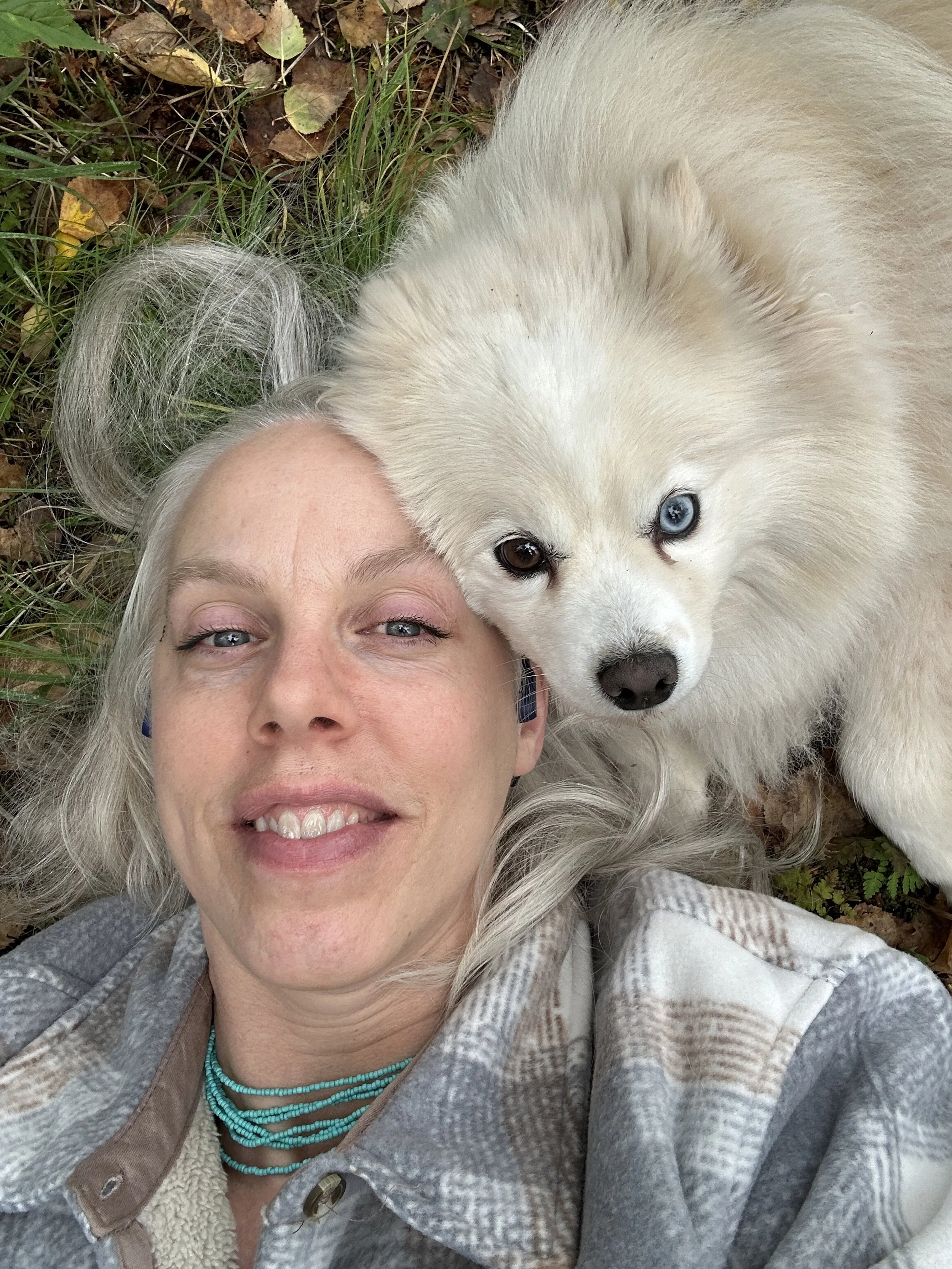 A woman with blond hair laying on the ground surrounded by leaves and grass, resting her head near a white dog with heterochromatic eyes (one blue and one brown).