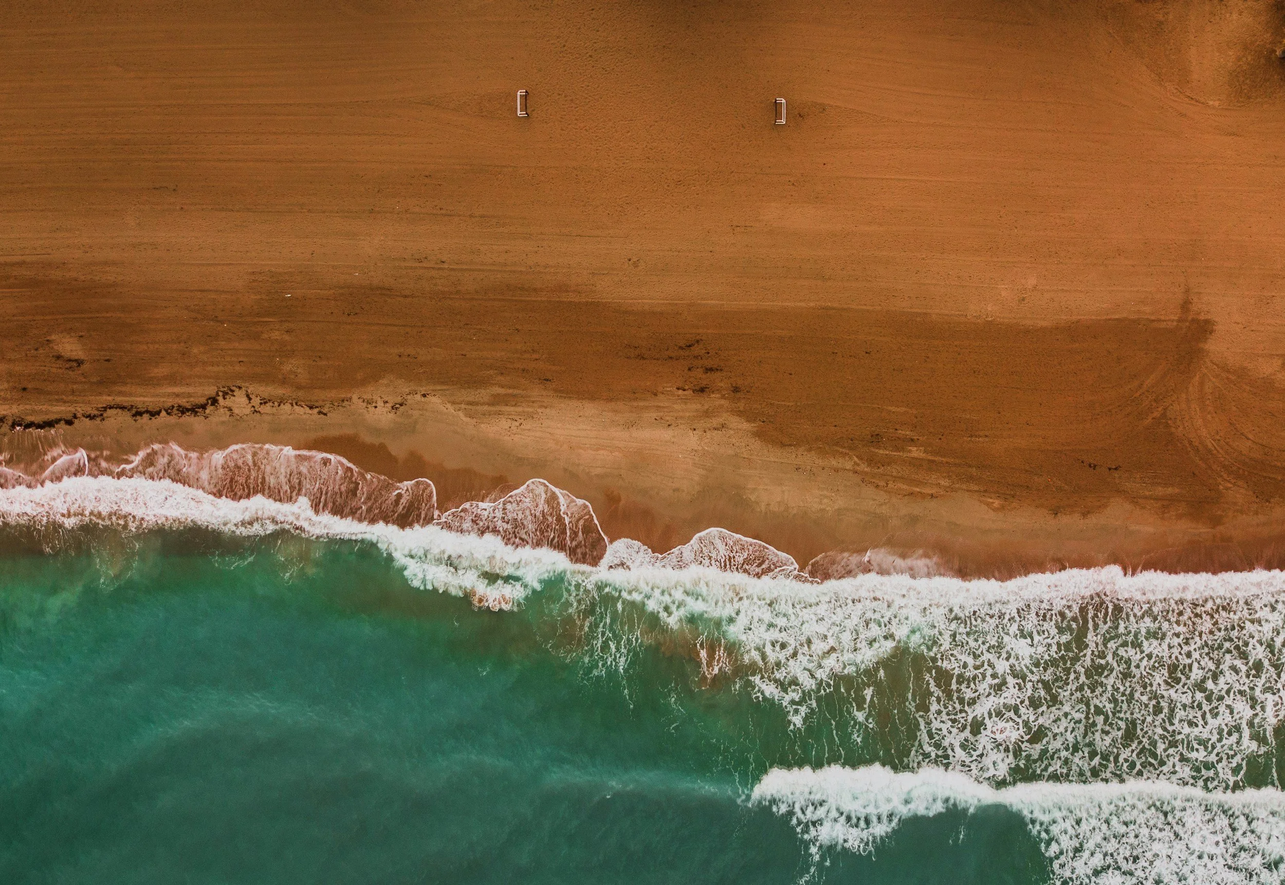 An aerial view of a sandy beach with ocean waves crashing onto the shore, showing green and white foam.