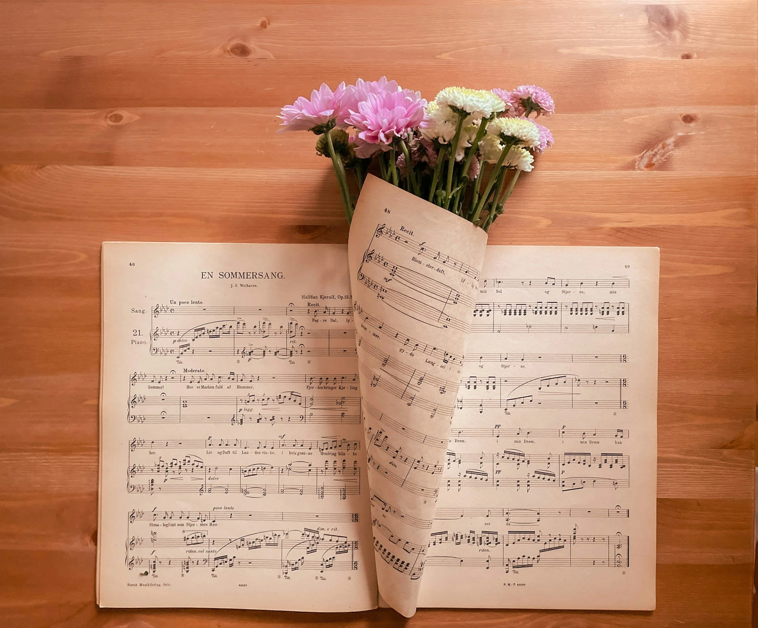 Open sheet music book on a wooden surface with a bouquet of pink and white flowers resting on top.