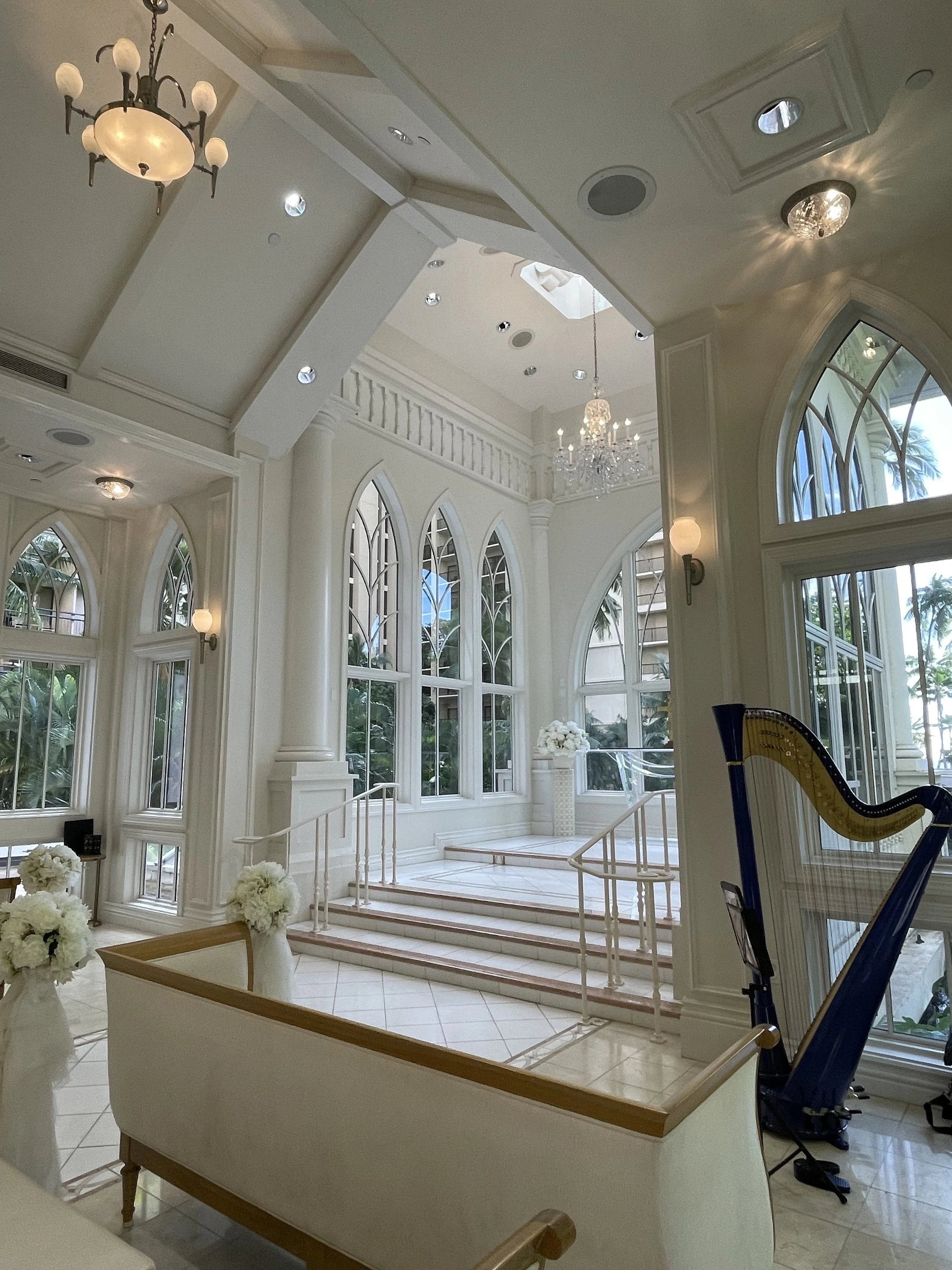 Elegant interior with white walls, large arched windows, a chandelier, and a harp near the window.