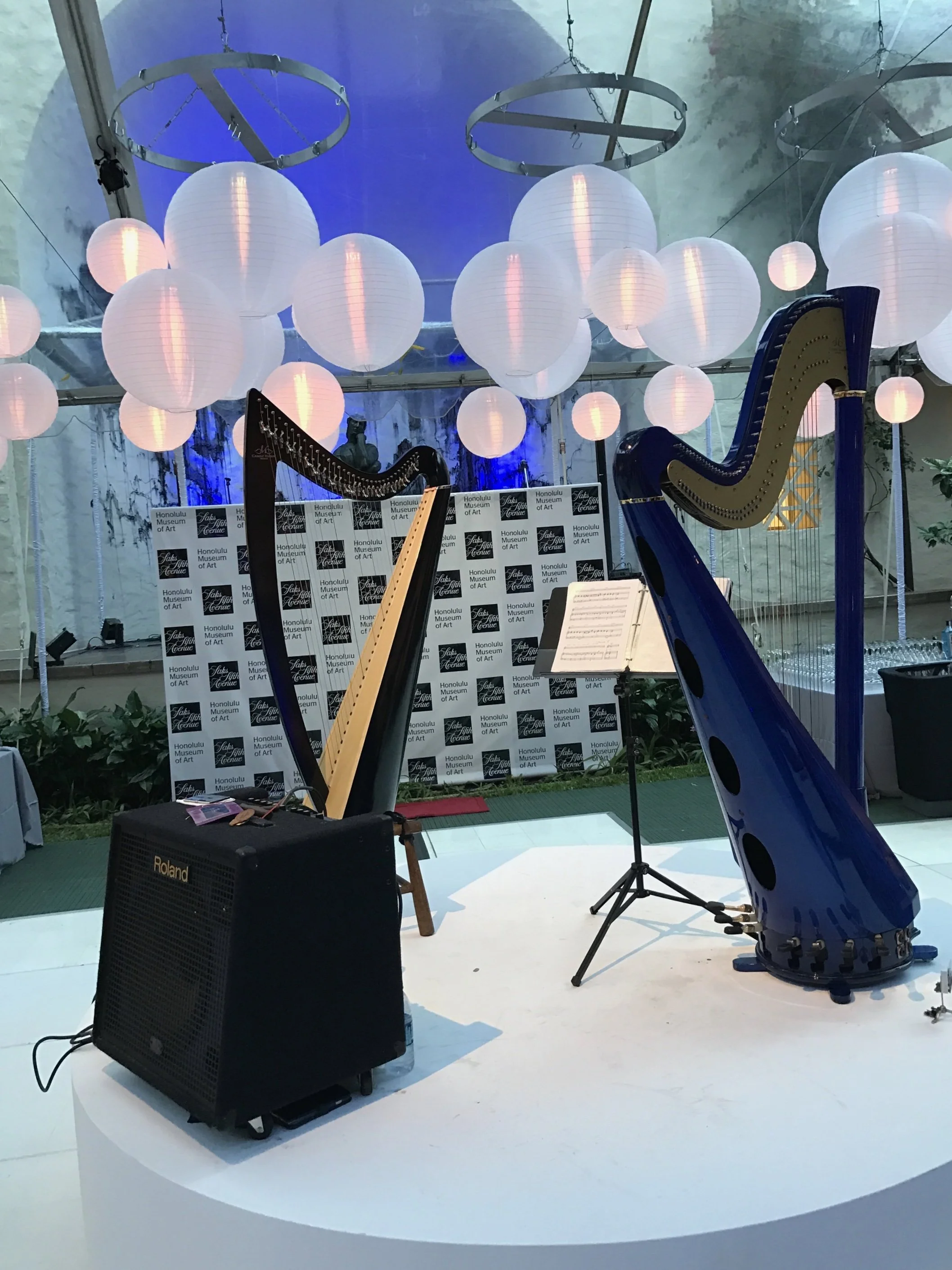 Two harps, one black and one blue, set up on a white stage with sheet music and a music stand. Decorative hanging paper lanterns and a backdrop with the Honolulu Museum of Art's logo.