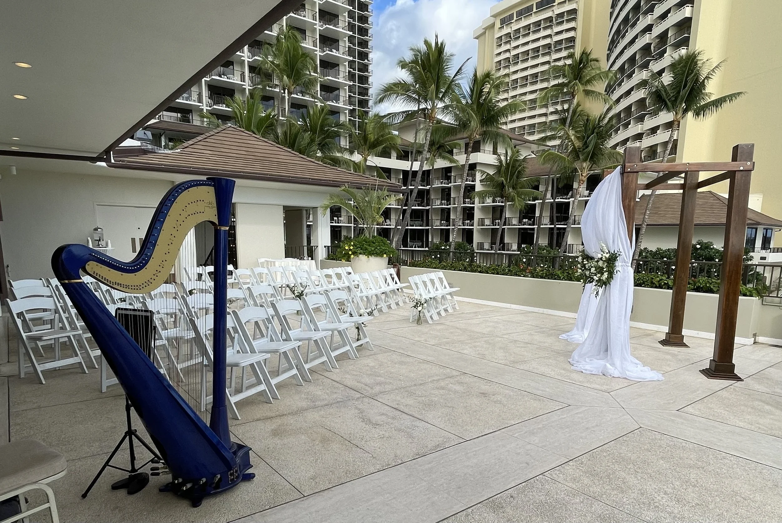 Outdoor wedding setup with a wooden arch decorated with white fabric and flowers, rows of white chairs, and a blue harp, with a background of tall apartment buildings, palm trees, and a cloudy sky.