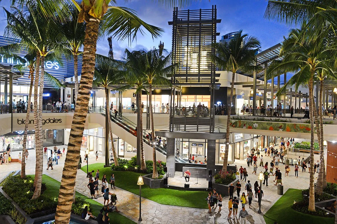 Outdoor shopping mall with palm trees, multiple levels, and people walking around, some seated near a small stage with a microphone in an open plaza area.