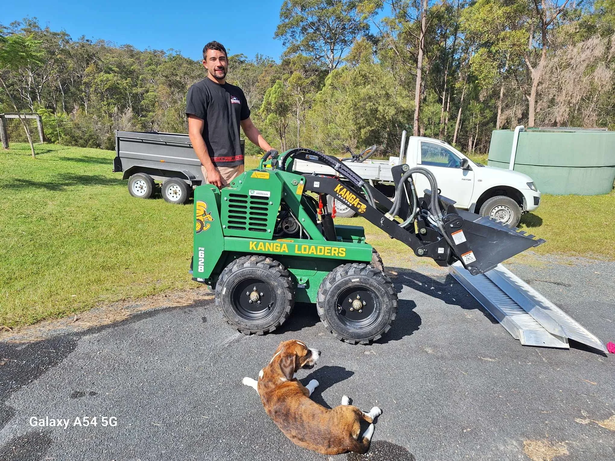 A man standing next to a green Kanga Loaders mini loader with a dog lying on the pavement and an artificial lawn in Canberra in front of it on a sunny day with trees and vehicles in the background.
