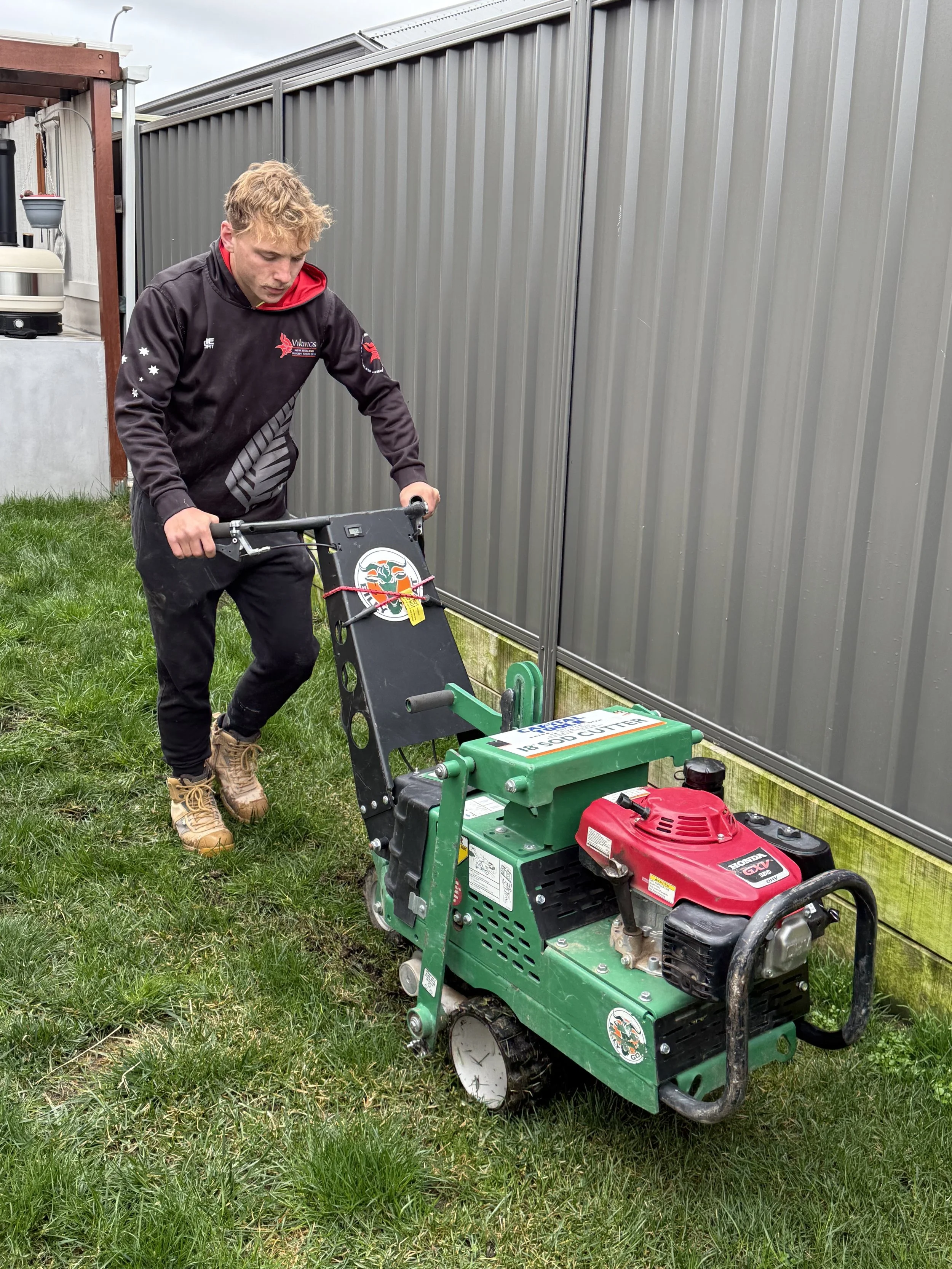 A person is laying bricks to create a walkway beside a brick house, with tools and construction materials around on a soil bed before the fake grass is placed in Canberra.