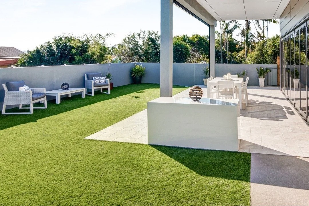 An image of the best fake grass in Canberra with a modern outdoor patio with green artificial lawn, white patio furniture including chairs and a table, decorative vases, potted plants, and a glass wall; surrounded by trees and clear sky.