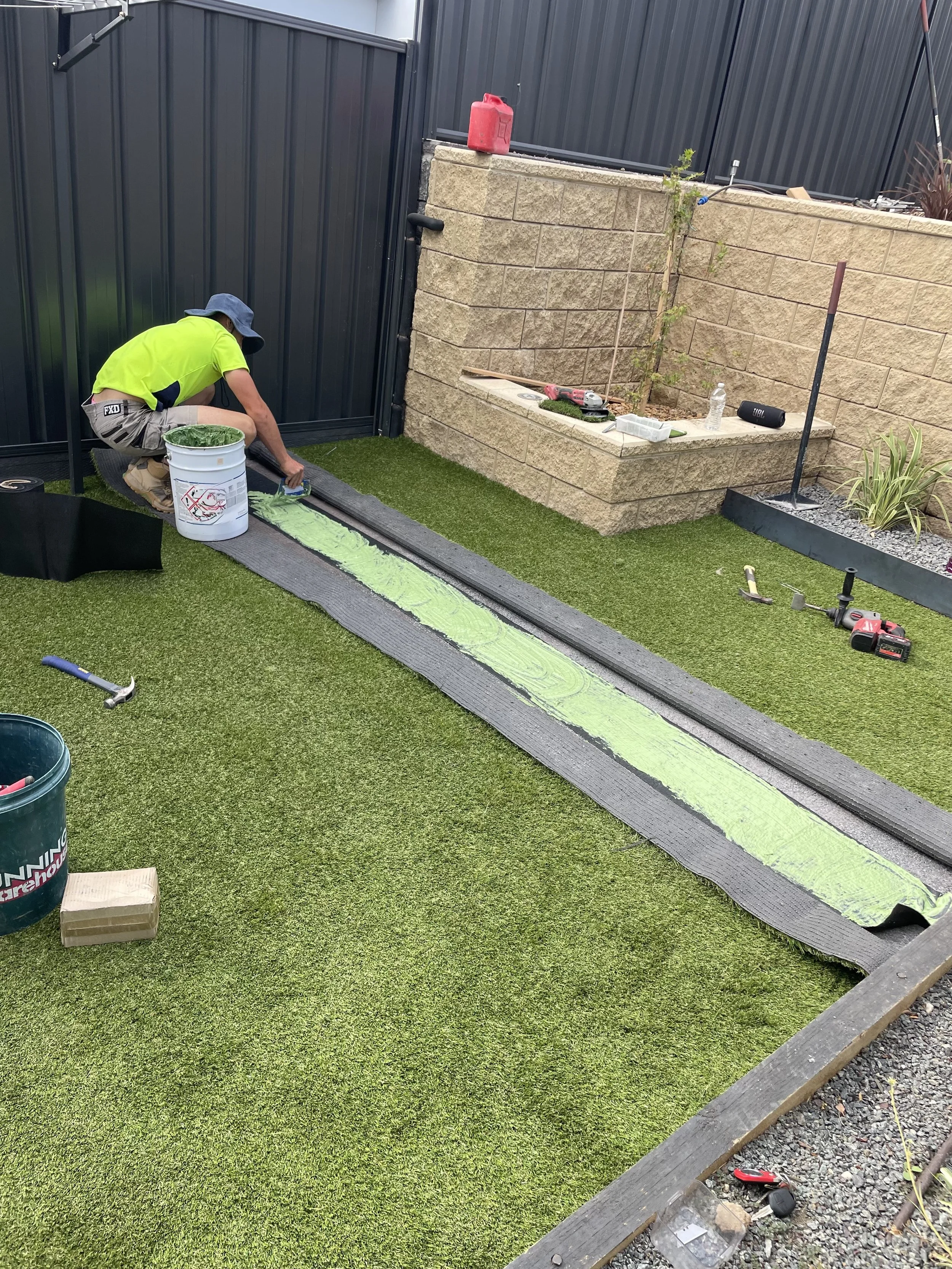 A person in a bright yellow shirt and wide-brimmed hat is installing a border around a small lawn, using black edging and a green adhesive strip. There are tools and supplies scattered around, including a bucket, a hammer, and power tools, with a partially built brick wall