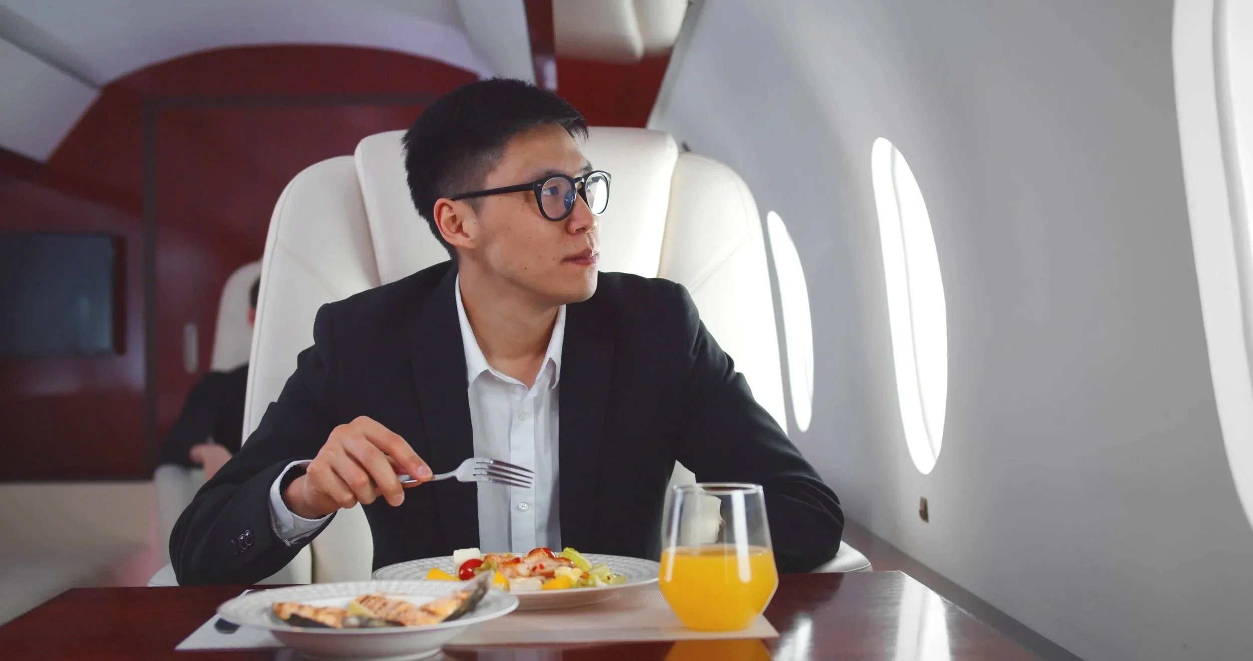 Man in black suit and glasses eating breakfast on airplane, with a plate of food, a glass of orange juice, and airplane windows in the background.