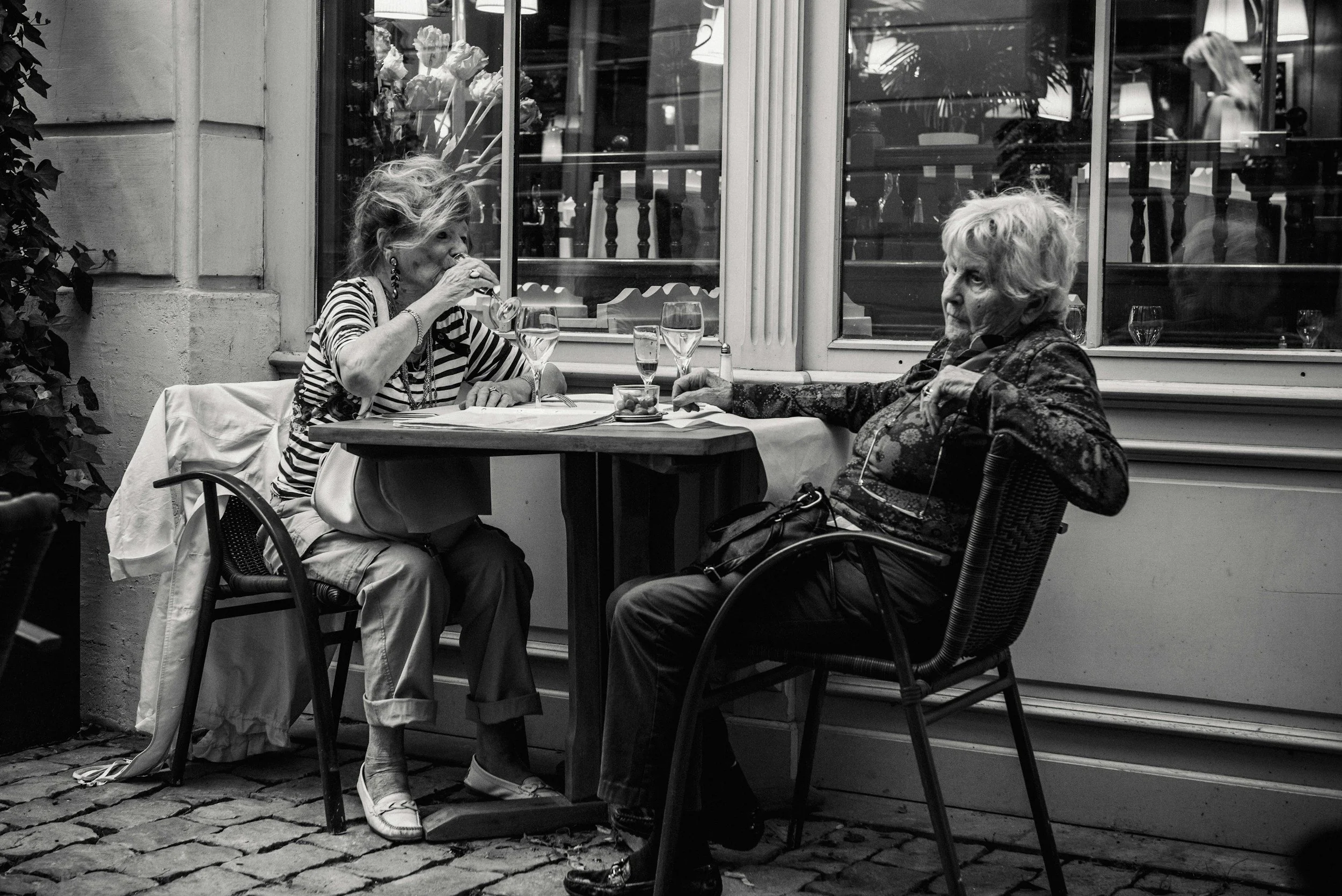 Two elderly women sit at a table outside a café, engaging in conversation. One woman is drinking from a glass, and the other is looking at her. The background shows large windows with reflections and a person inside.