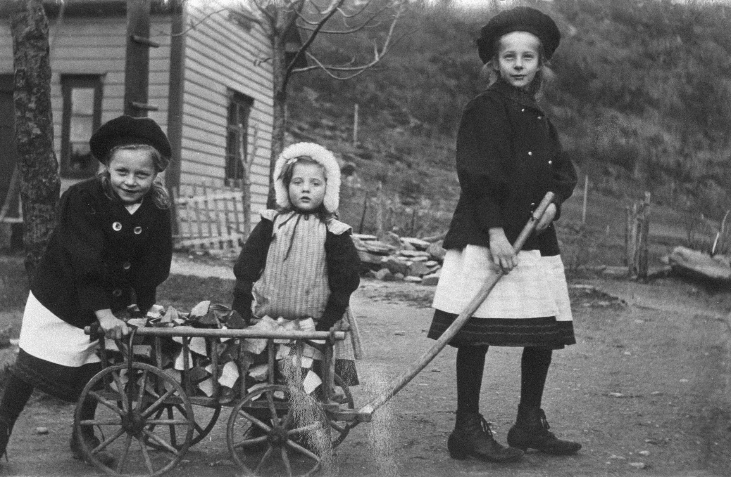 Three young girls outdoors near a house, two pulling a small wagon filled with objects and the third standing beside them holding a stick, all dressed in older-style clothing and hats, in a rural setting.