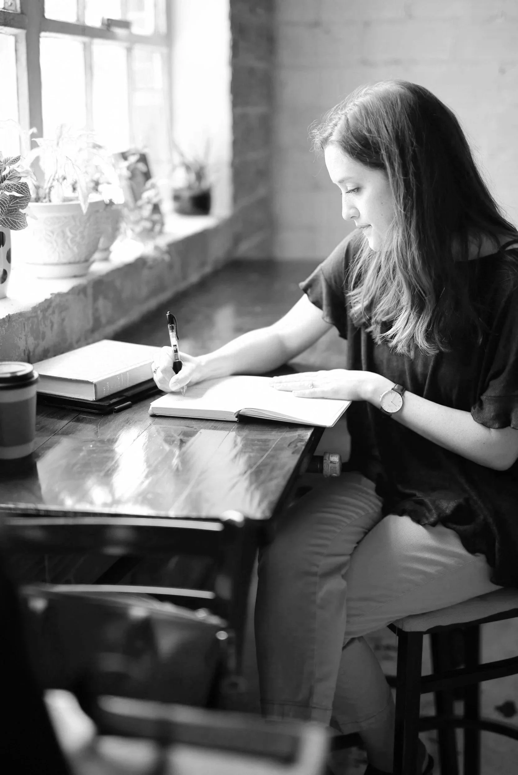 A young woman sitting at a wooden table, writing in an open notebook with a pen. There are a few books and a coffee cup on the table. She is near a window with potted plants, in a cozy, well-lit room.