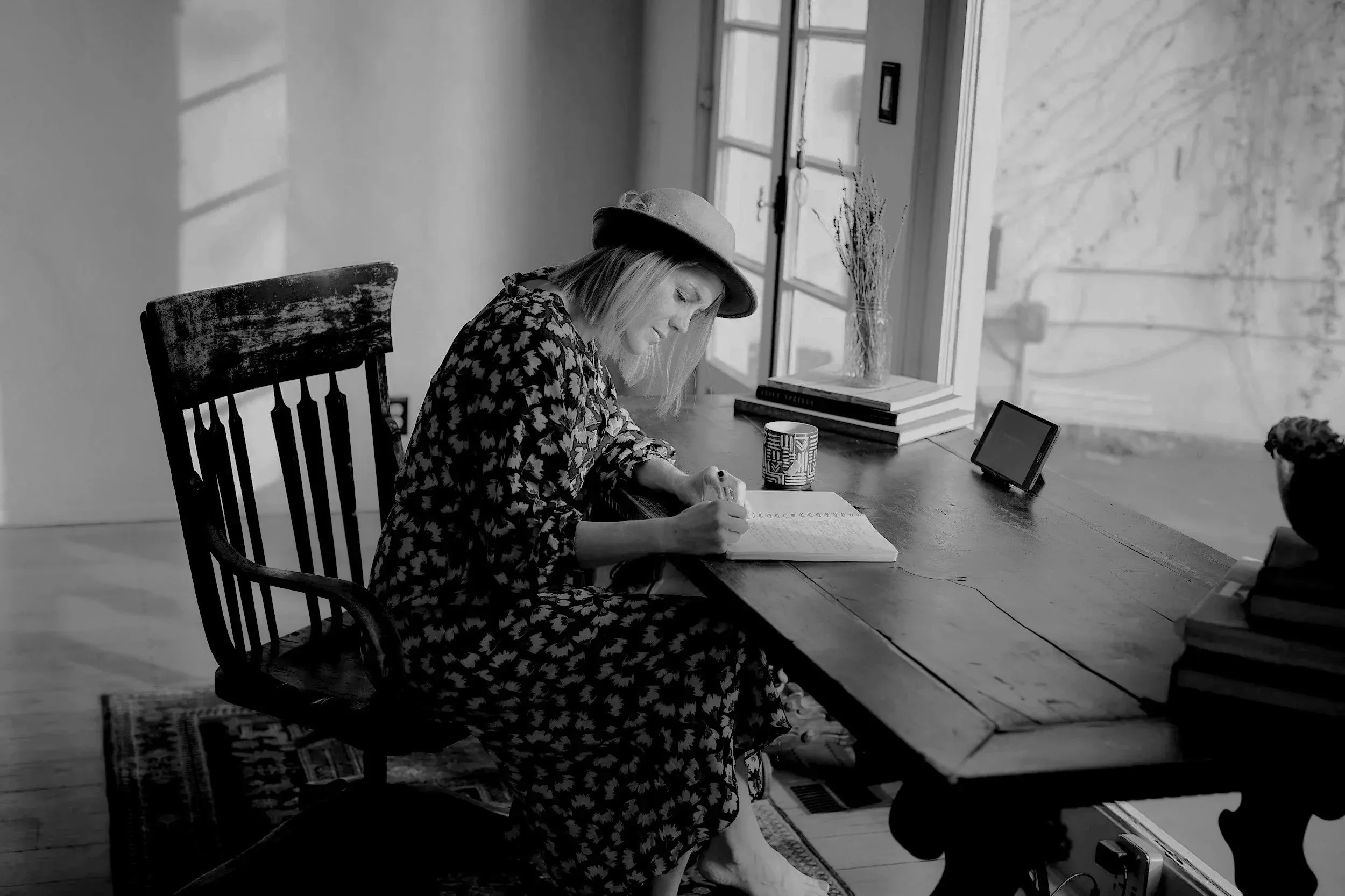 A woman sitting at a wooden desk writing in a notebook, wearing a hat and patterned dress, beside a window with a vase and books, with a small tablet on the desk.