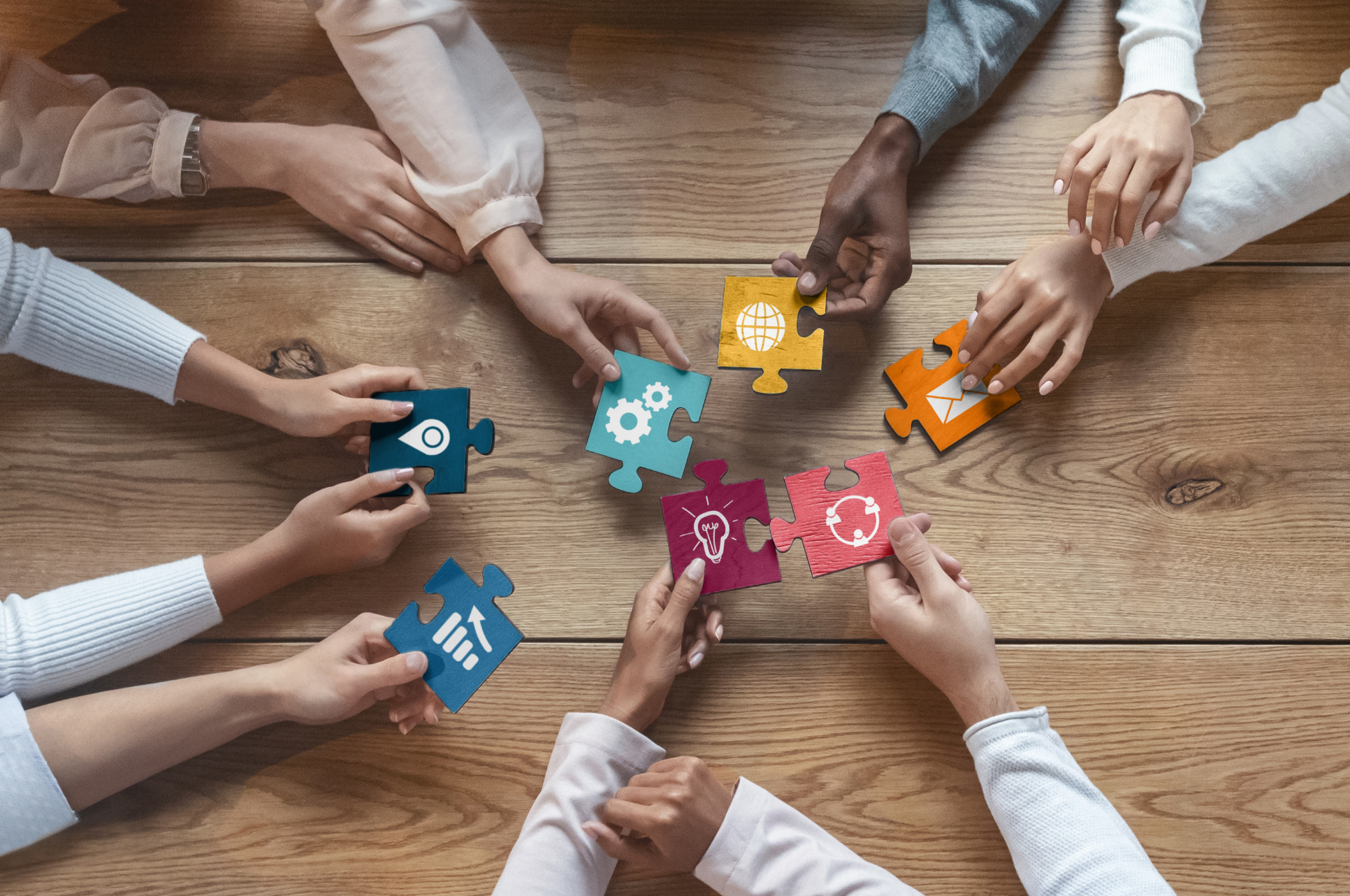 Multiple hands holding colorful puzzle pieces with icons representing global communication, gears, email, light bulb, headset, and handshake, on a wooden table.