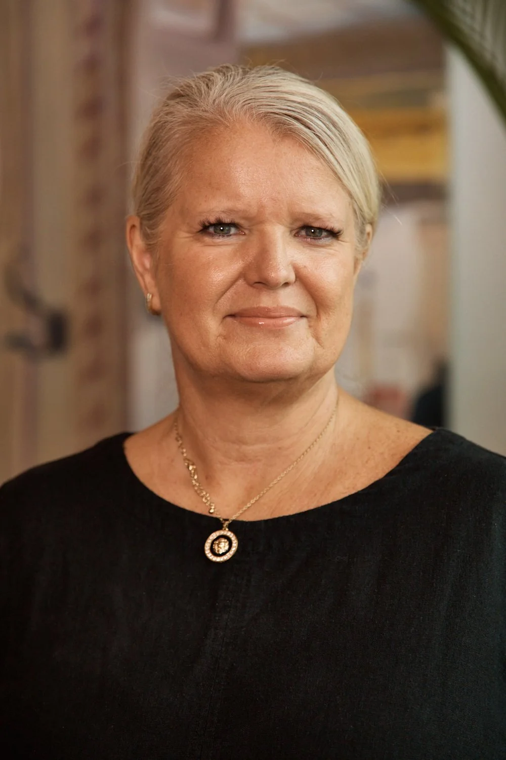 A middle-aged woman with short blonde hair, wearing a black top and a gold necklace with a circular pendant, standing indoors.
