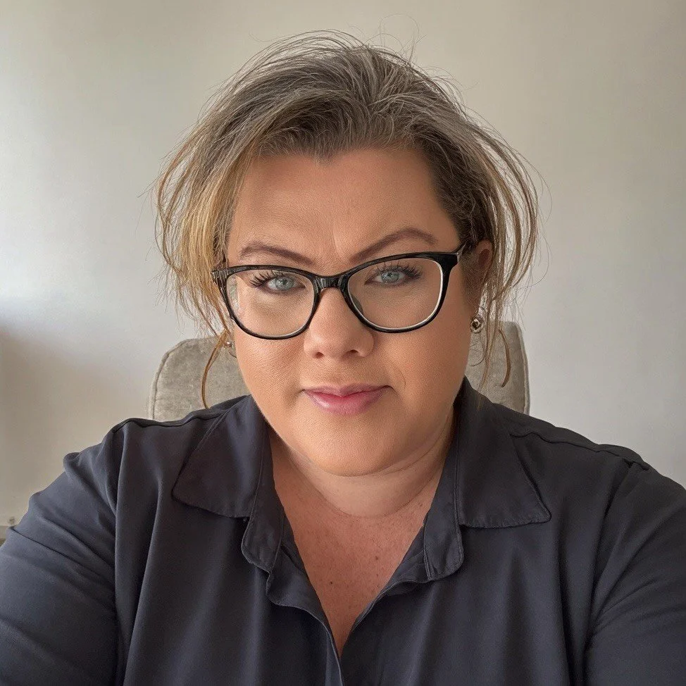 A woman with short, tousled blonde hair, glasses, and a black collared shirt, sitting indoors against a plain, light-colored wall.