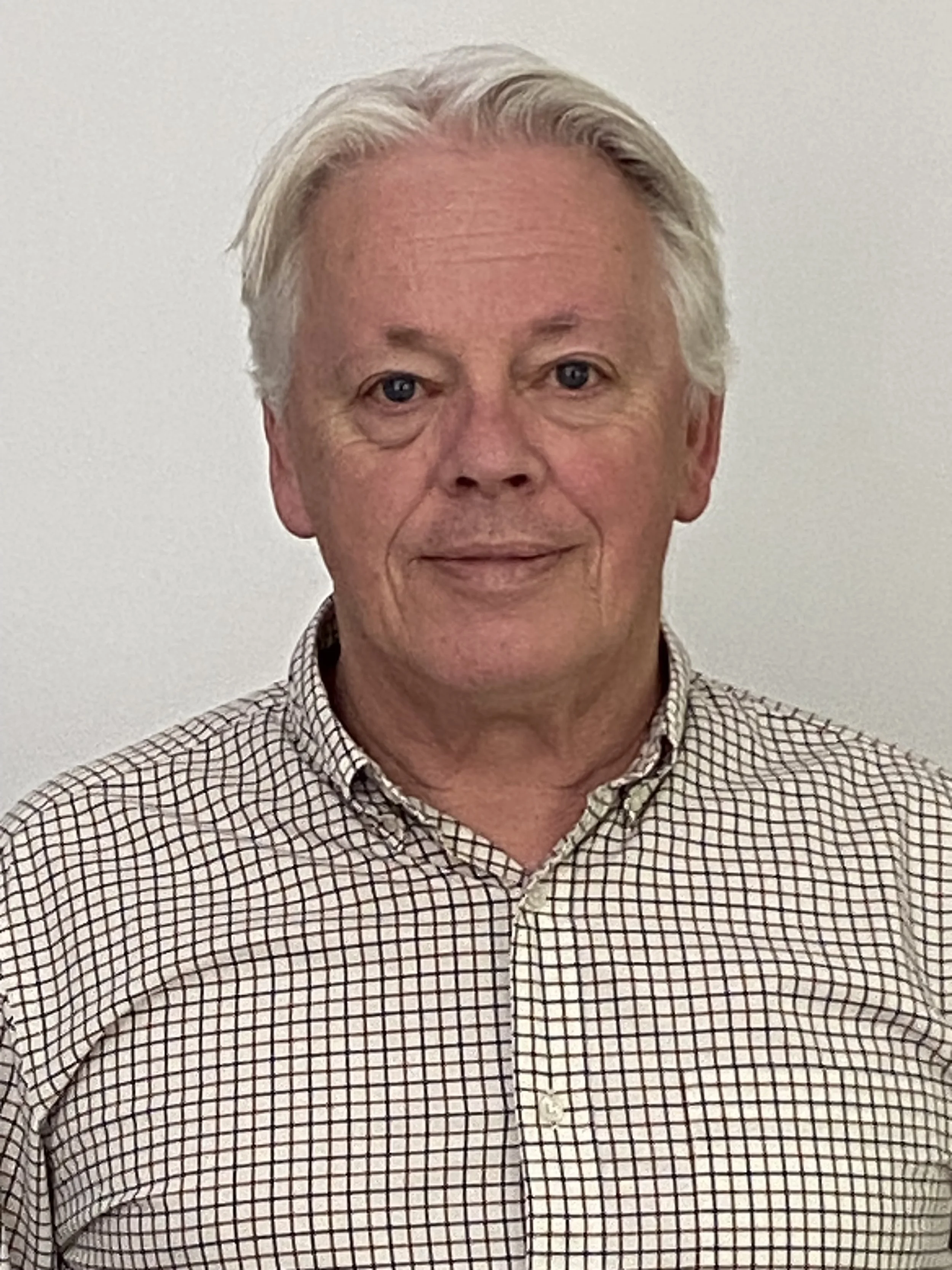 Headshot of an elderly man with gray hair, wearing a checkered button-up shirt, standing against a plain white wall.
