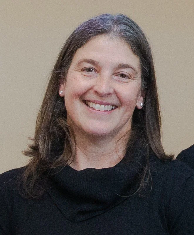 A woman with shoulder-length brown hair, smiling, wearing pearl earrings and a black turtleneck sweater, standing against a beige background.
