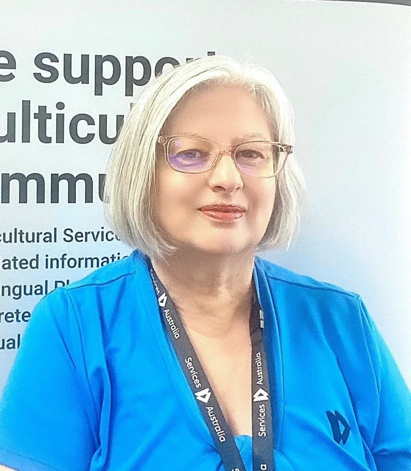 A woman with short, gray hair and glasses, wearing a blue shirt and lanyard, standing in front of a sign with partially visible text.