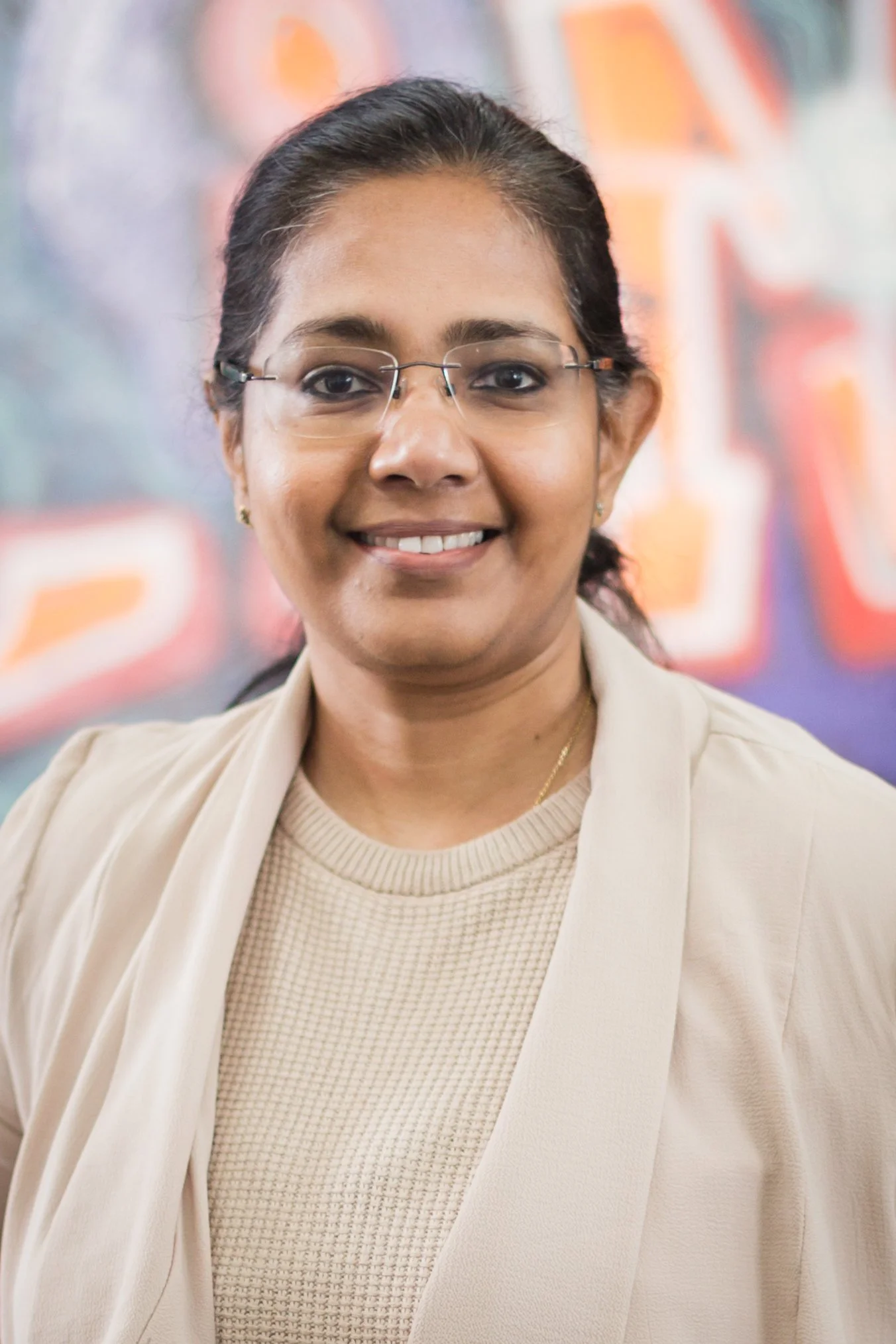 A woman wearing glasses and a light-colored blazer, smiling at the camera. Background has colorful blurred graffiti.