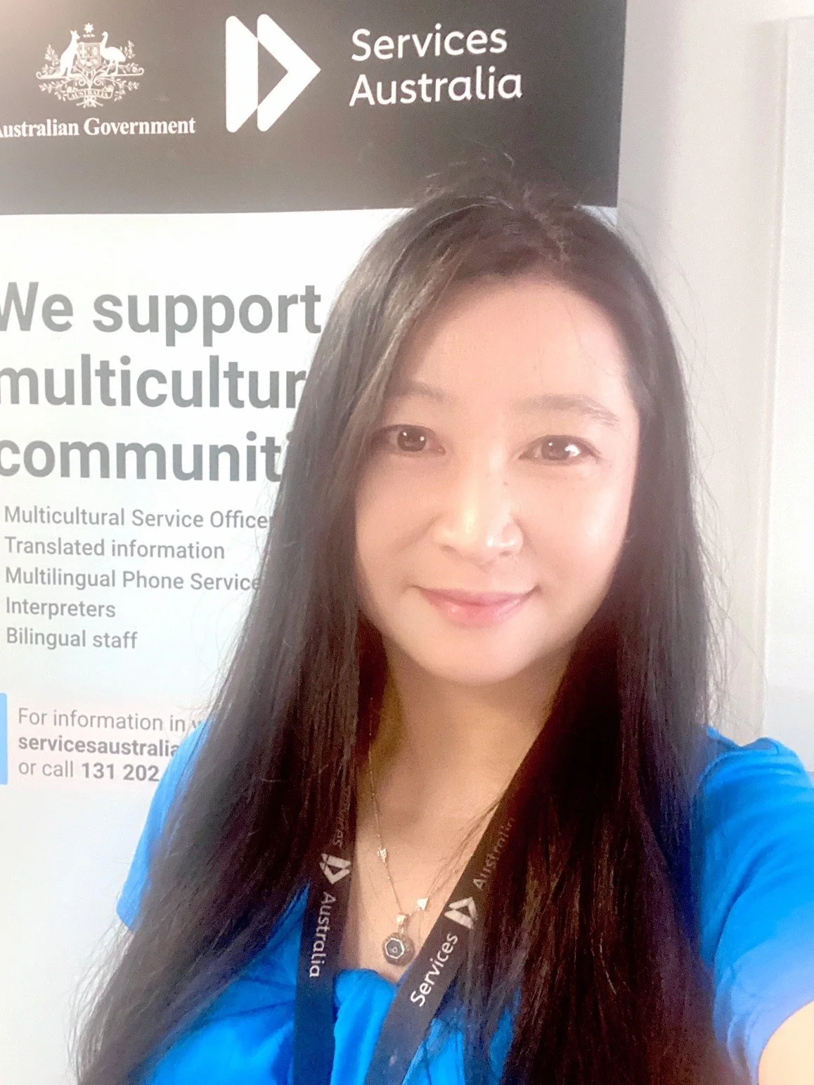 A woman with long dark hair wearing a blue shirt and a black lanyard taking a selfie in front of a sign for Services Australia at the Australian Government.