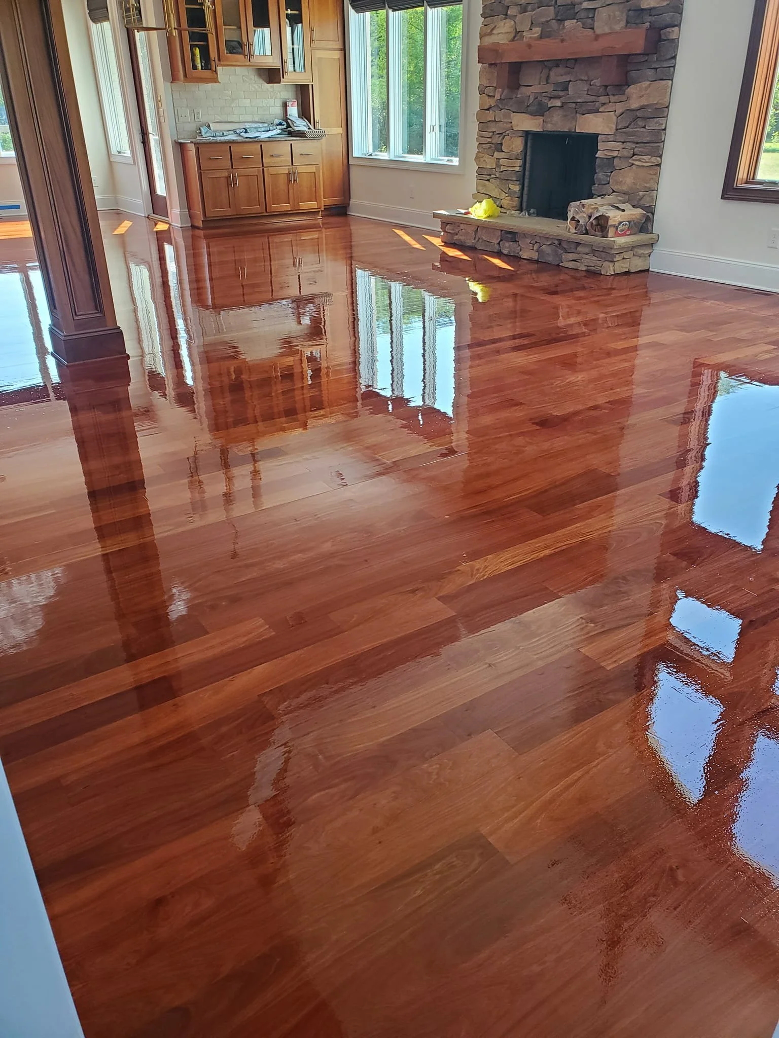 Polished hardwood floor in a living room with stone fireplace, large windows, and wood cabinetry above a countertop.