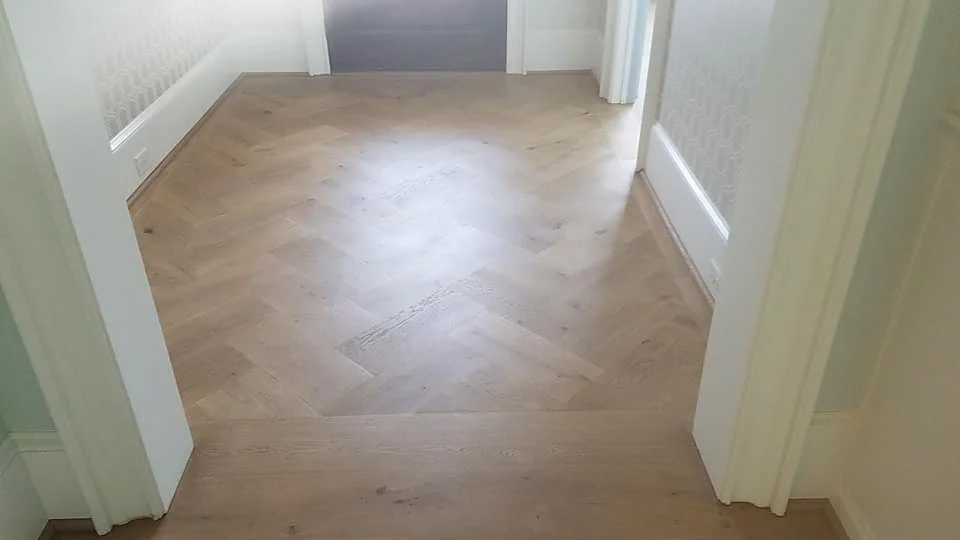 Empty hallway with wooden herringbone patterned floor and white walls.
