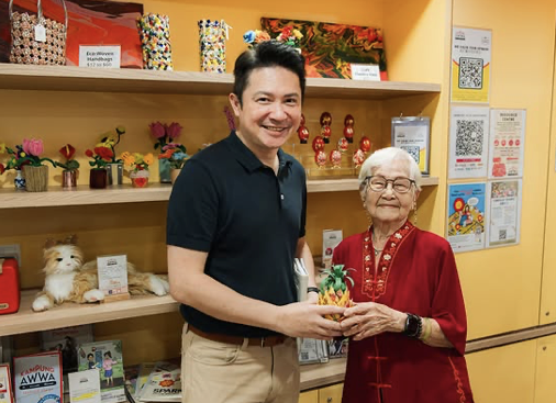 A man and an elderly woman smiling and holding a small decorative item in a colorful store with shelves of toys, books, and decorations.