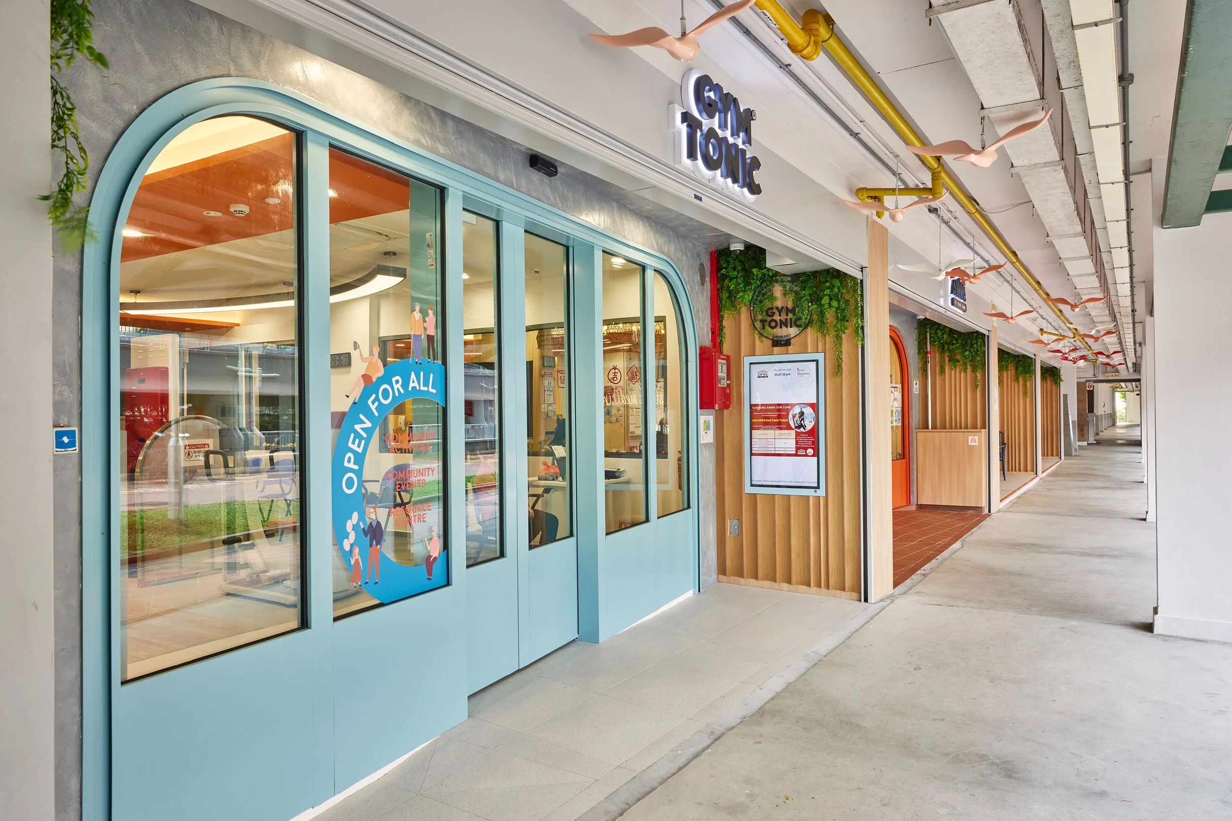 Entrance to a fitness center named Gym Tonic with a blue door, signage, and a digital display outside, located in a shopping area with a wide walkway.